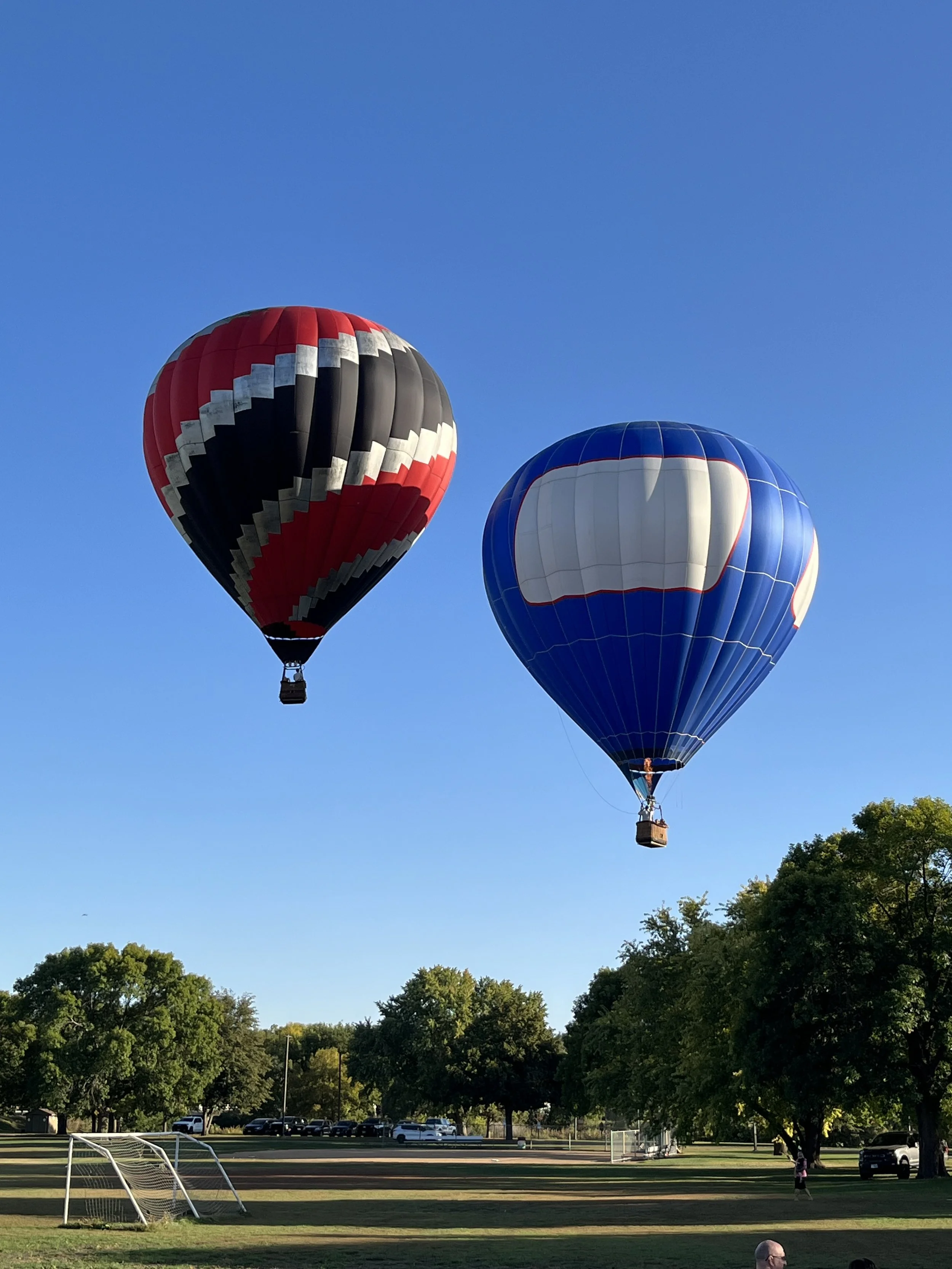 Two hot air balloons floating in the sky over a park with green trees and a soccer goal post.