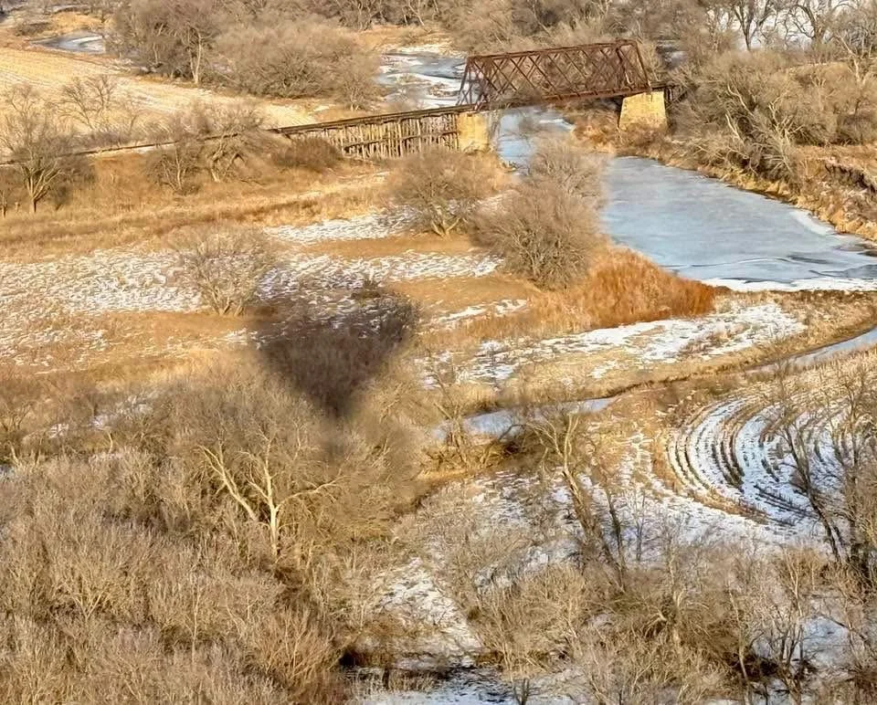 An aerial view of a river flowing through a brown, snowy landscape with leafless trees. An old metal bridge crosses the river in the background, and a shadow of a person is cast on the ground.