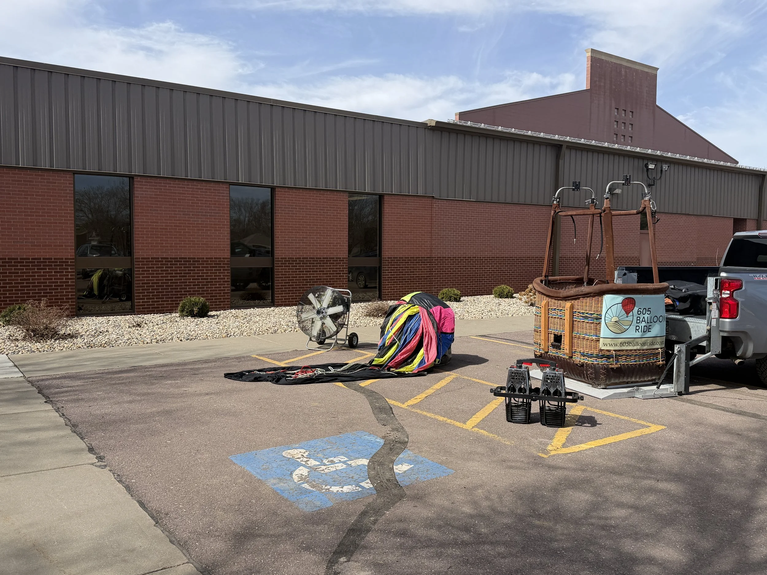 Hot air balloon basket with balloons and equipment in a parking lot next to a brick building, marked as a handicap space, with a fan and baskets nearby.