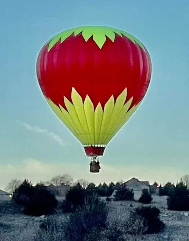 A colorful hot air balloon designed to look like a strawberry, with red and green sections, floating in the sky over a hilly landscape with trees and houses.