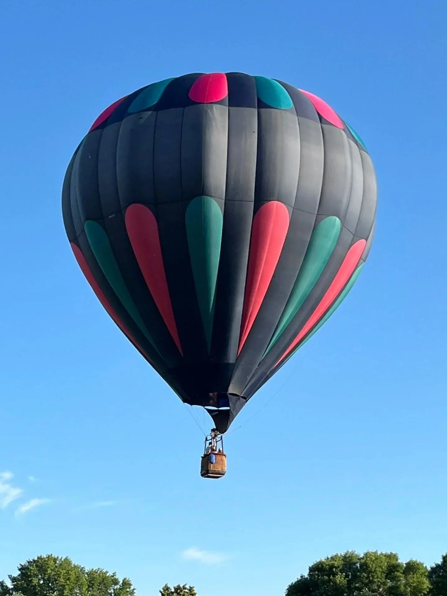 Colorful hot air balloon flying in a clear blue sky, with green trees visible at the bottom.