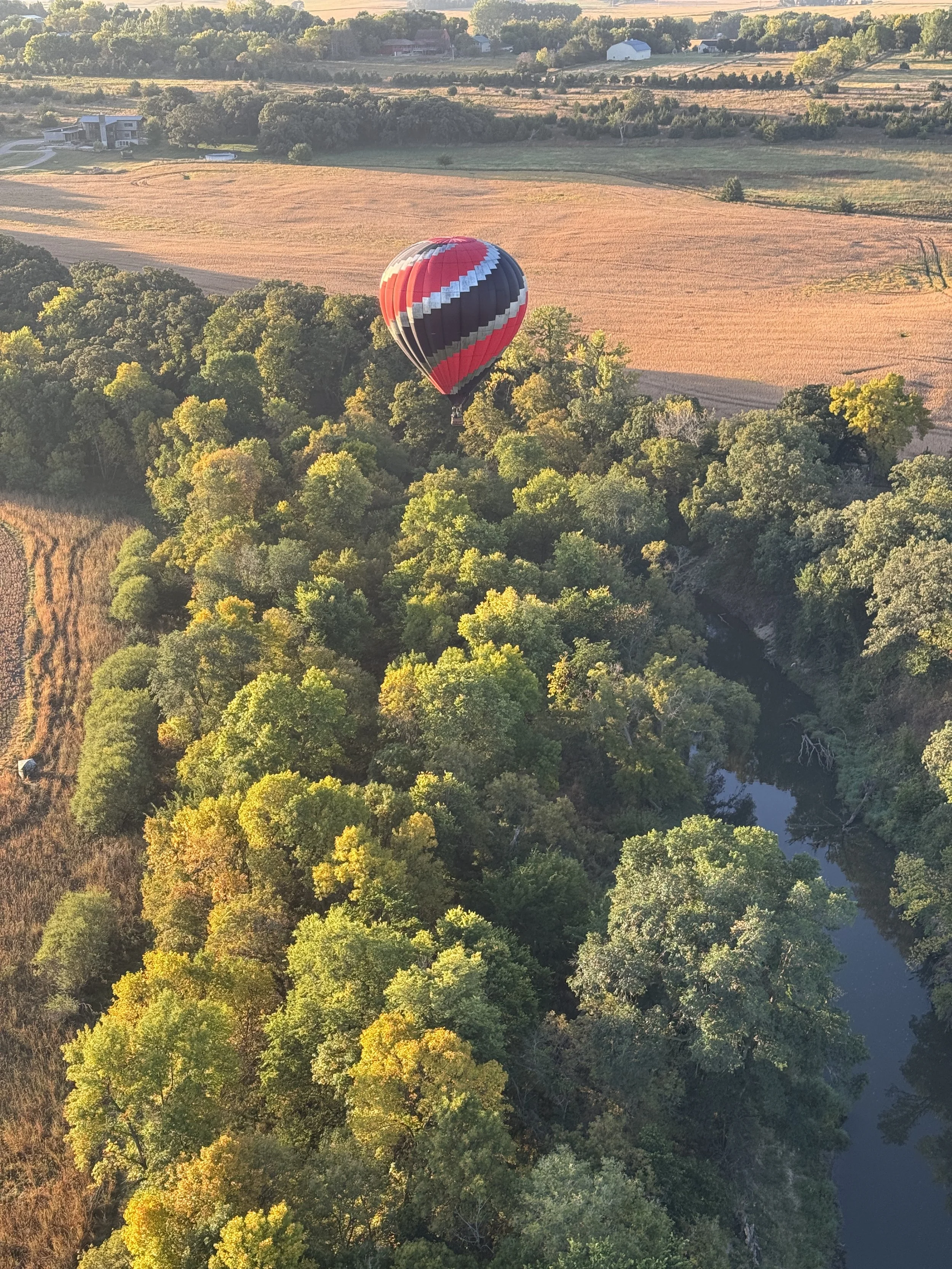 A hot air balloon with red, black, and white stripes flying over a green treetops near a river in a rural landscape.