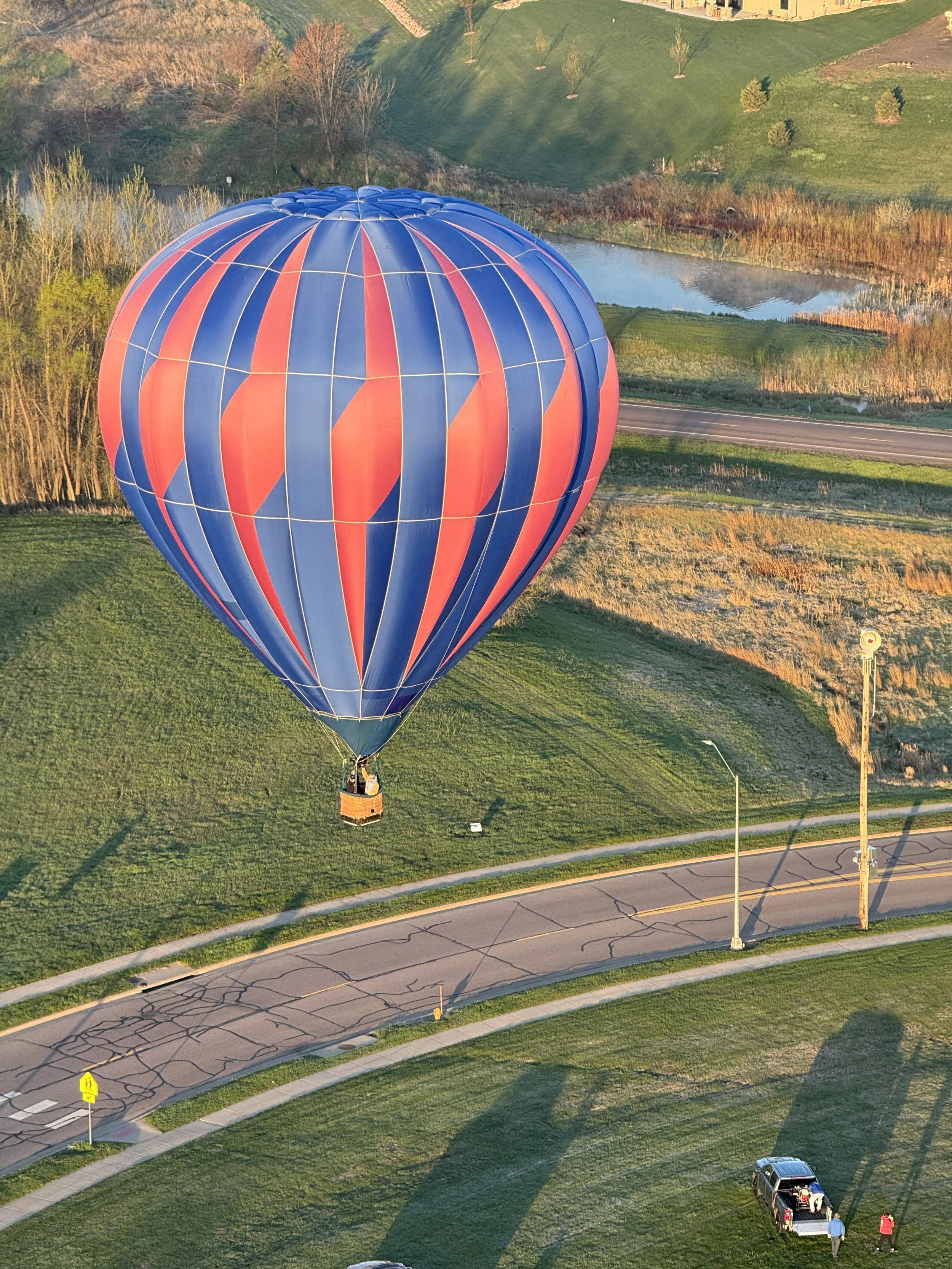 A hot air balloon with blue and red vertical stripes floating above a green park area, with a small pond and a road visible in the background.