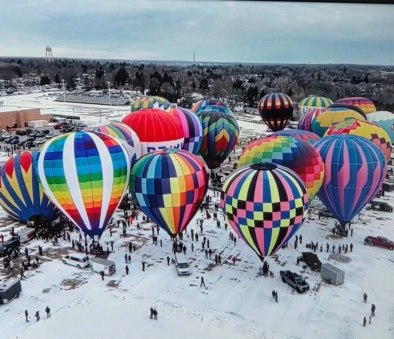 Hot air balloons with colorful patterns and designs in a snowy open area, with people and vehicles nearby, under a partly cloudy sky.