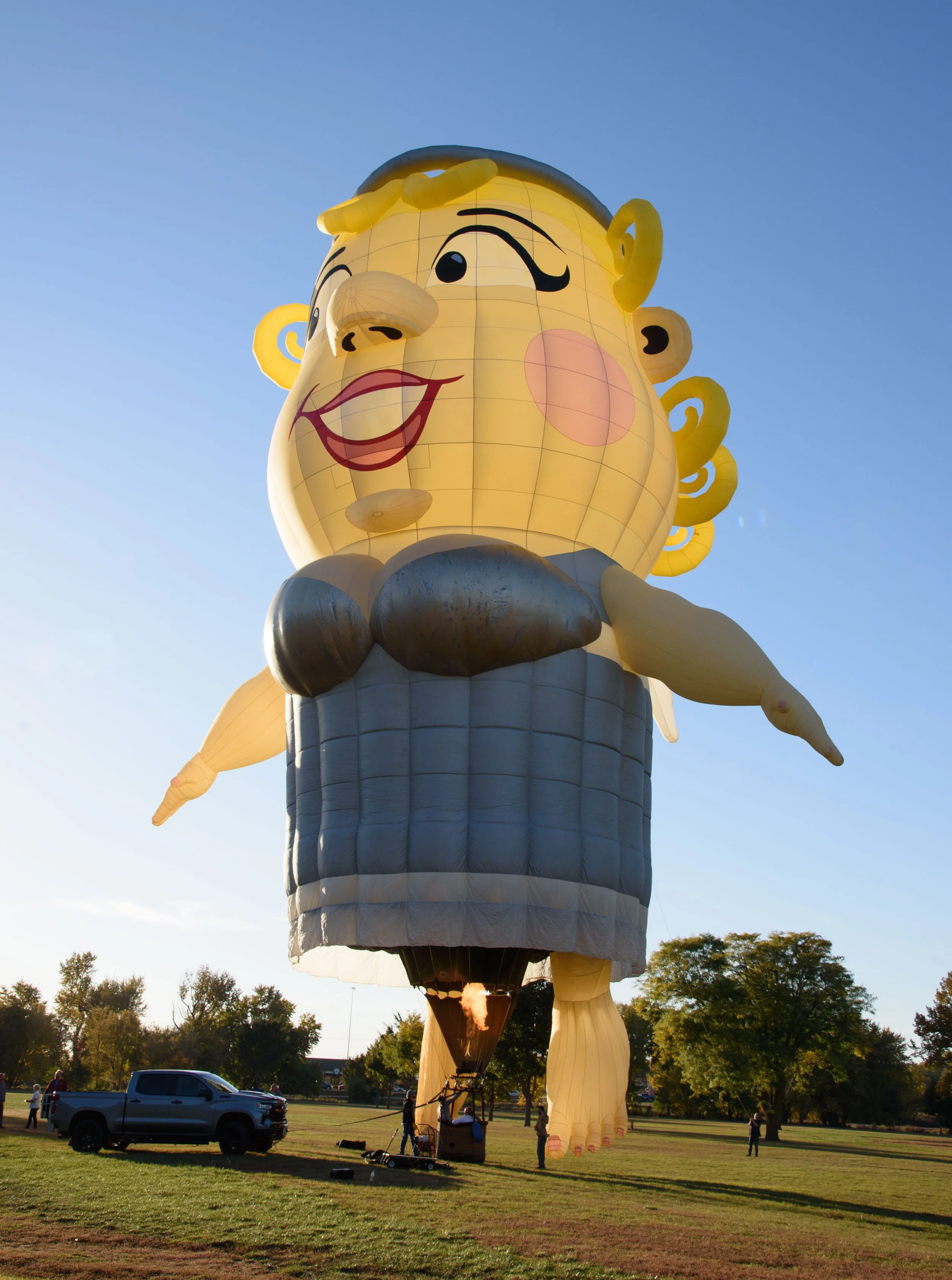 A large hot air balloon shaped like a cartoon woman with yellow hair, a pink cheek, and a big smile, standing on a field with trees in the background during sunset.