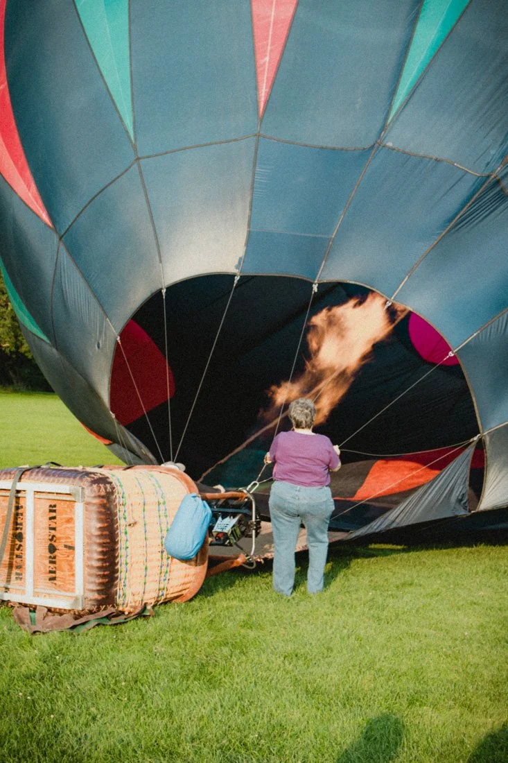 A hot air balloon is on the ground, partially deflated, with a woman standing in front of it. The balloon's fabric shows colorful patterns and is spread out on a grassy field, with the basket lying on its side near the woman.