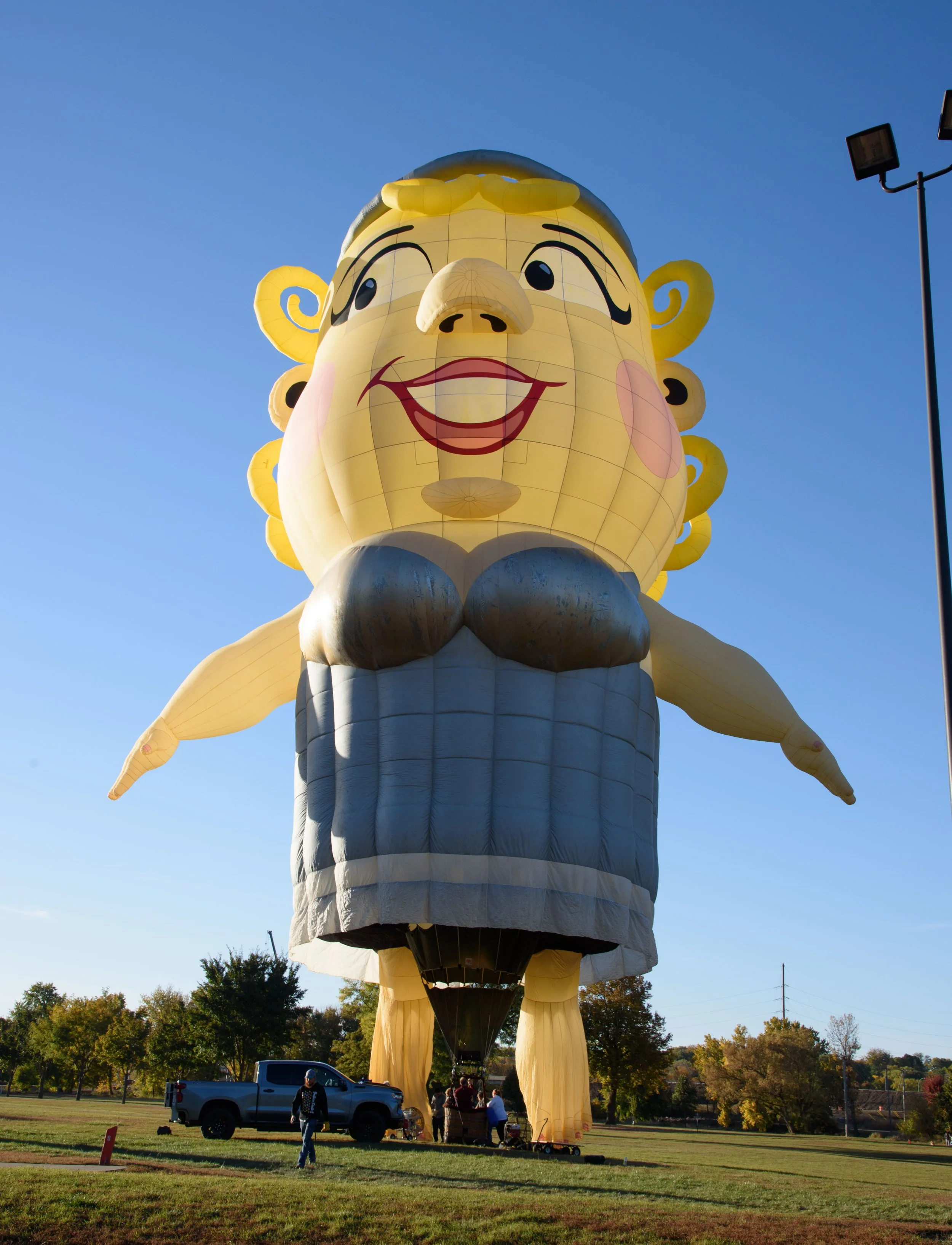 A giant hot air balloon shaped like a smiling woman with curly hair, a nose, and red lips, wearing a blue dress. People are seen near the balloon and a truck is parked nearby. The sky is clear and blue.