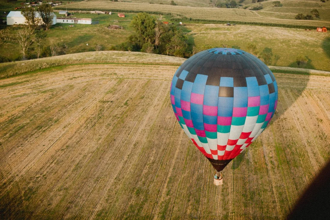 A colorful hot air balloon with a checkered pattern floating above a rural farmland landscape with fields, trees, and some buildings in the distance.