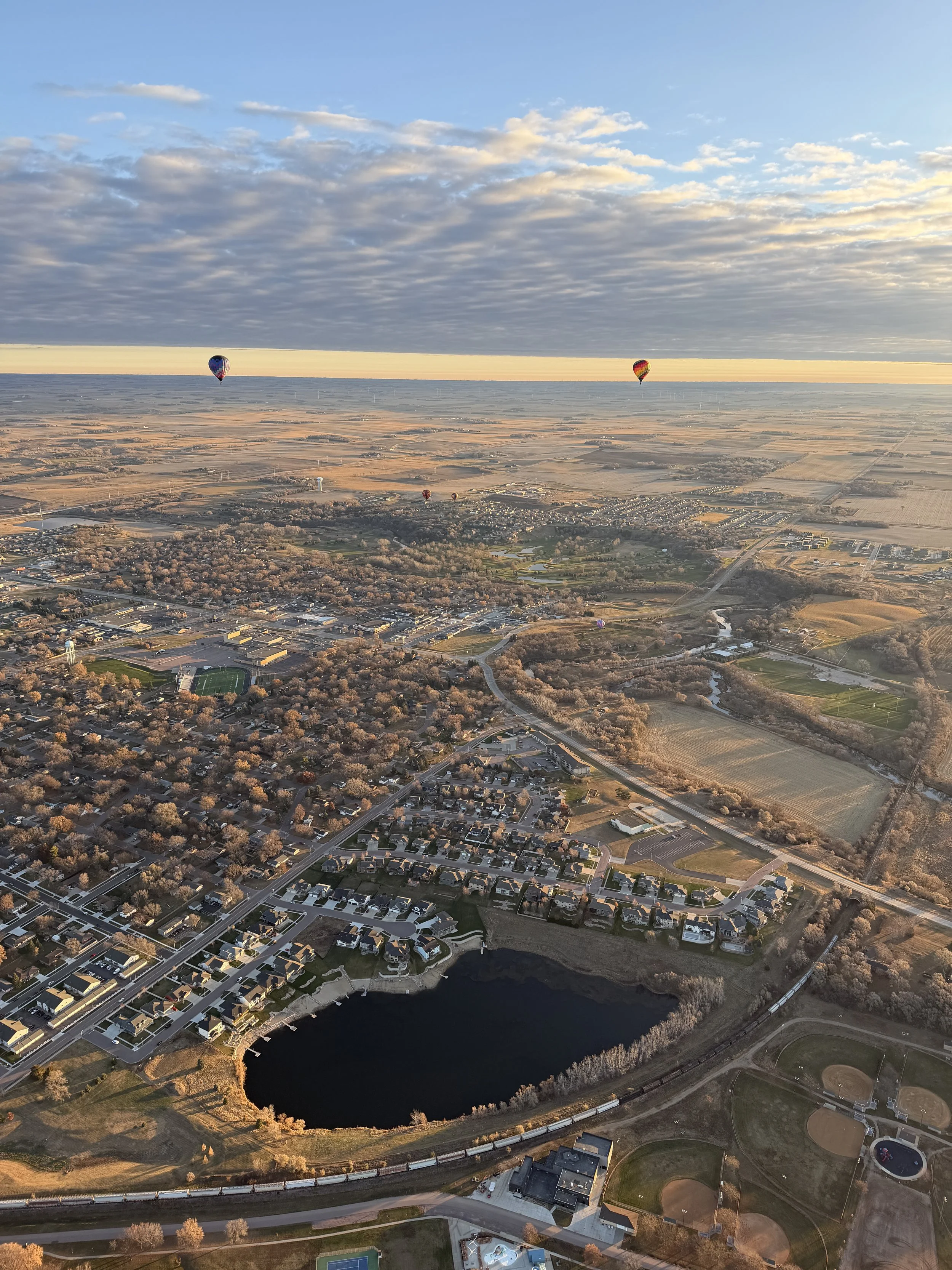 An aerial view of a suburban area with houses, roads, a lake, and a park, with five hot air balloons floating in the sky during sunset and a partly cloudy sky.