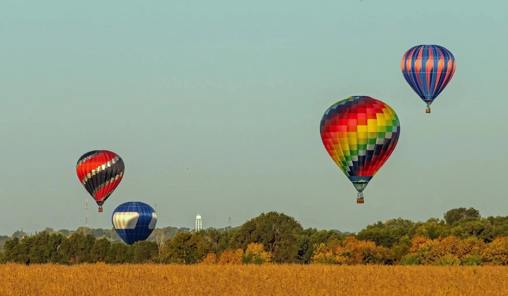 Five colorful hot air balloons floating over a green and orange autumn landscape.