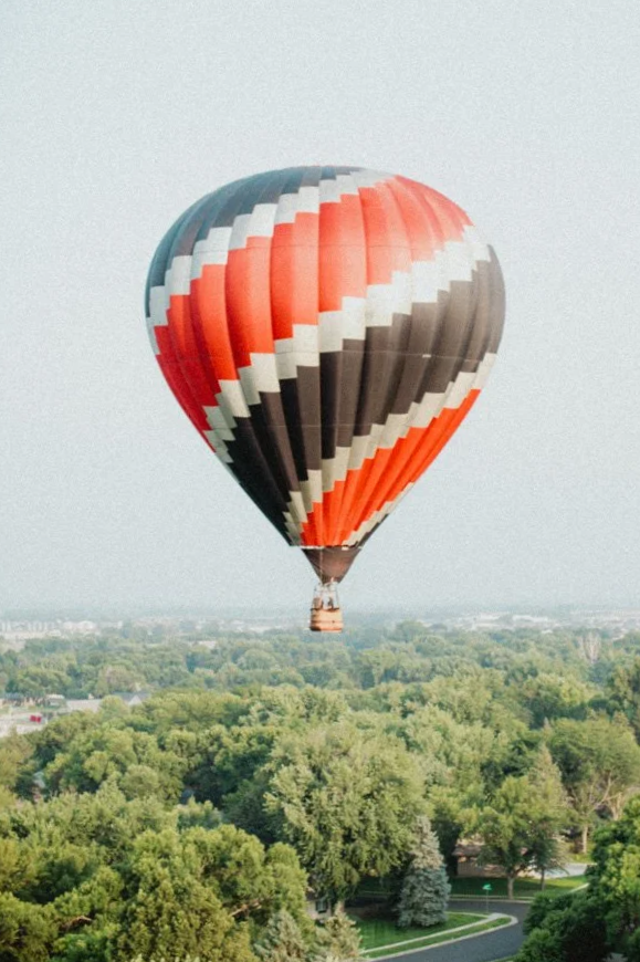 Colorful hot air balloon floating above green trees and a suburban area.