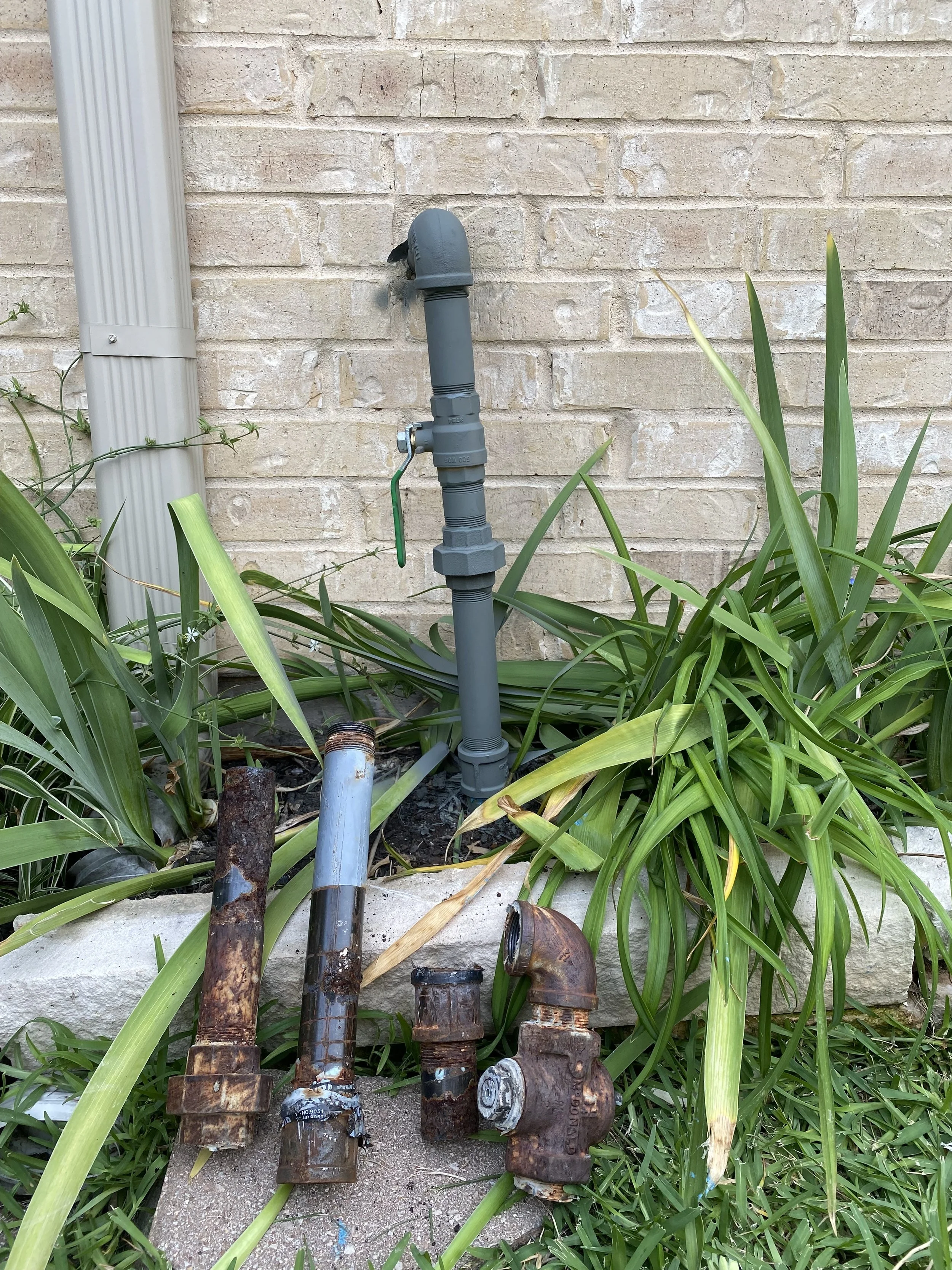 A garden with a green-leafed plant near a brick wall. There are rusty and old pipe fittings and a grey outdoor faucet with a shut-off lever.