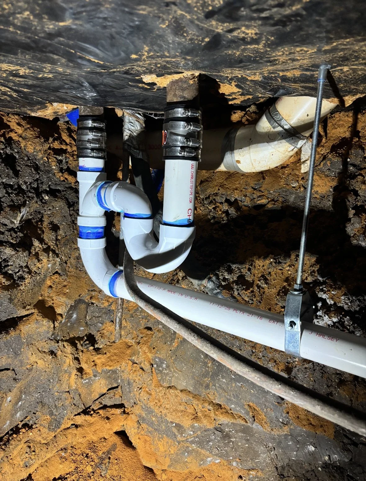 Underground plumbing system with PVC pipes, fittings, and insulation, secured with pipe straps, installed in a rocky, dirt-filled space.