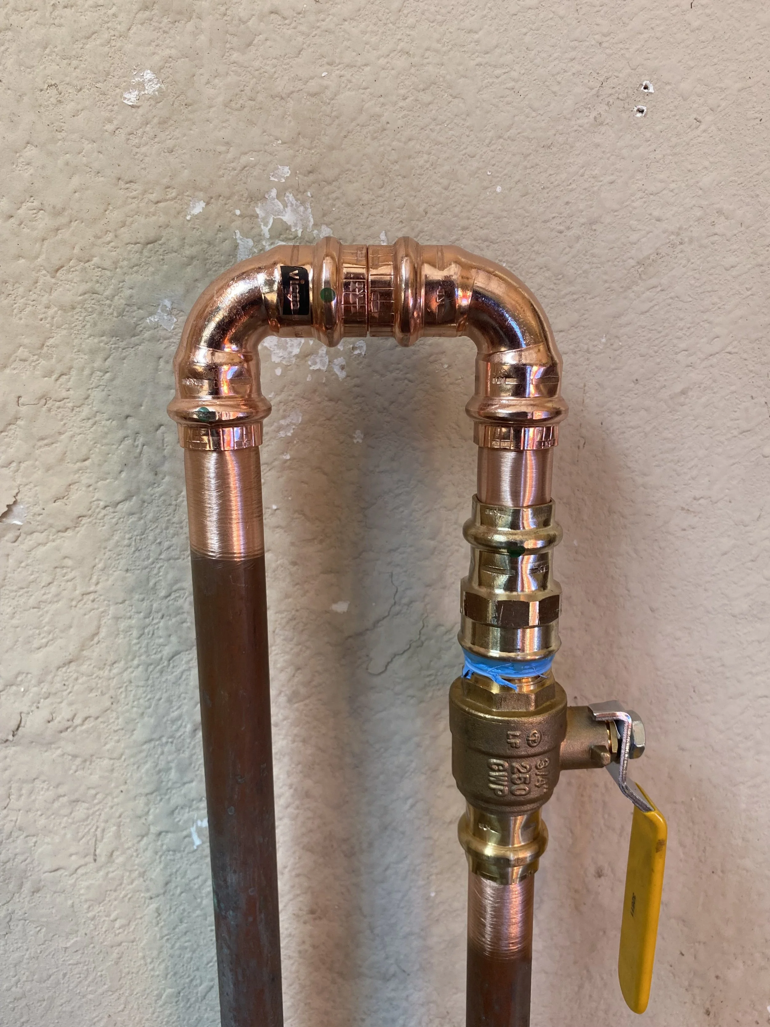Copper plumbing pipes with a brass shut-off valve and yellow handle against a beige textured wall.