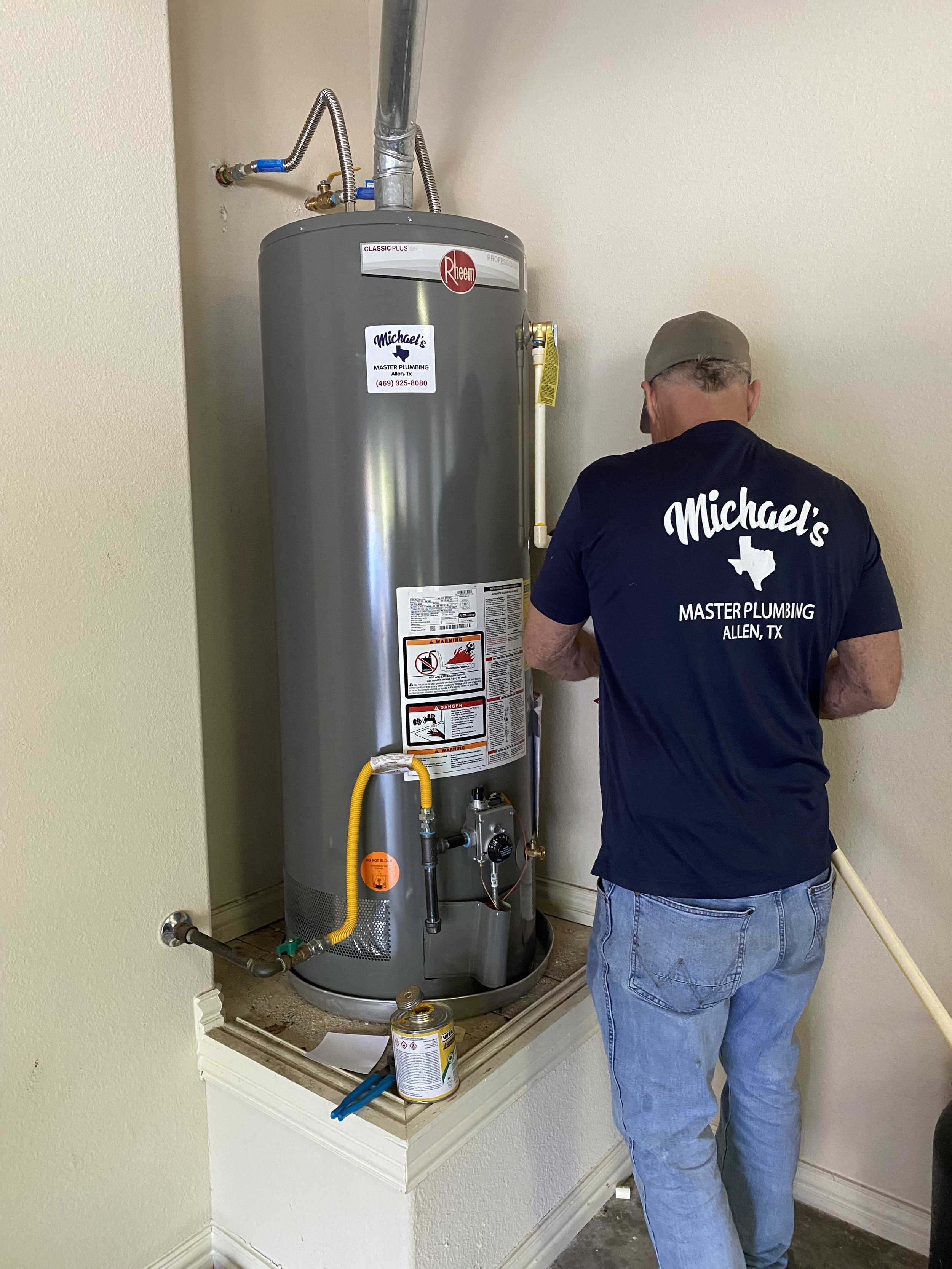 A plumber working on a water heater in a residential utility area. The water heater is grey with various labels and a yellow gas line connected to it. The plumber is wearing a navy blue t-shirt with 'Michael's Master Plumbing, Allen, TX' printed on t