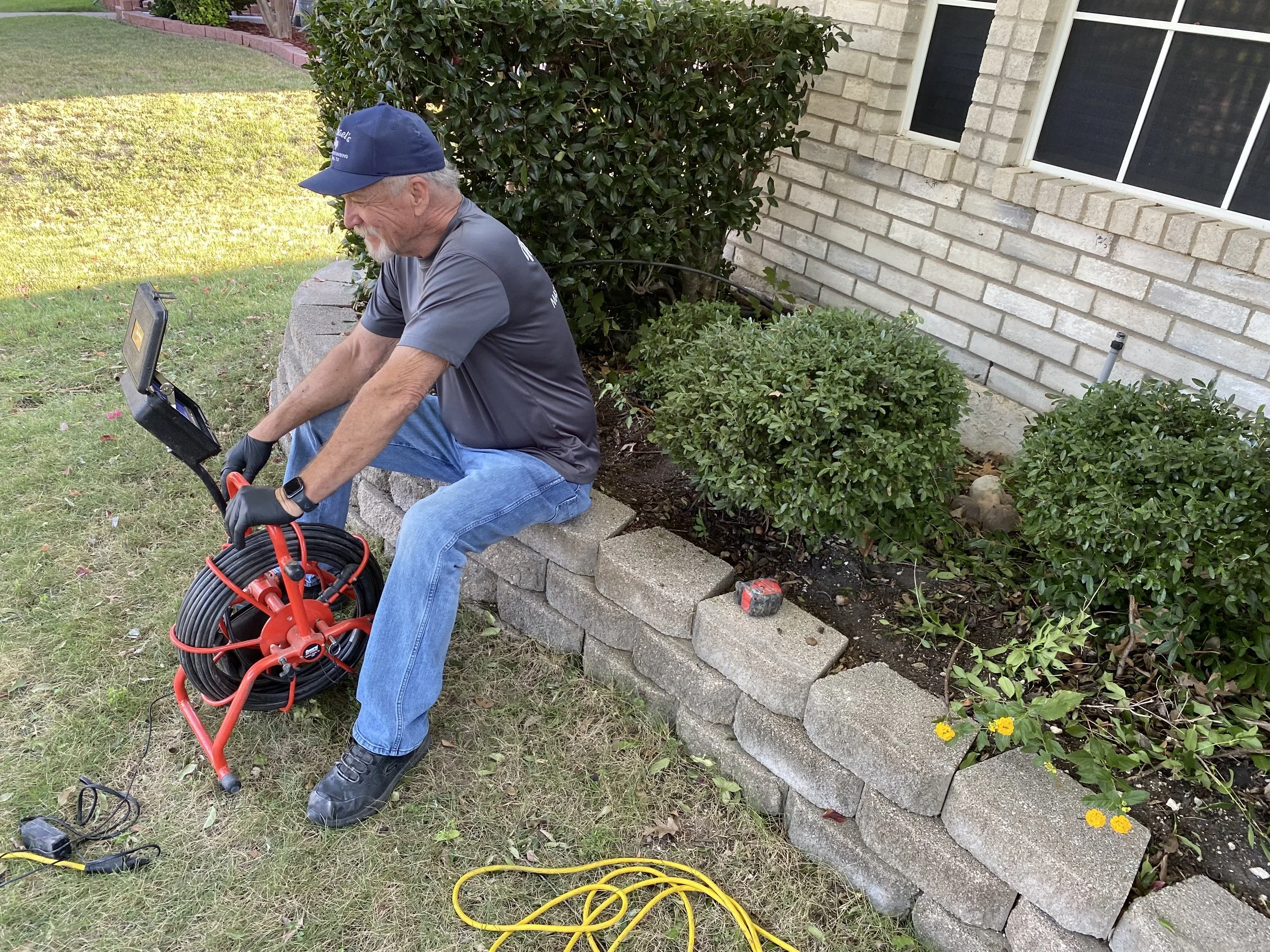 A man wearing a navy cap, gray t-shirt, and blue jeans is sitting on a low brick wall, handling a red extension cord reel. He is outdoors in a yard with grass, bushes, and a brick house with windows.