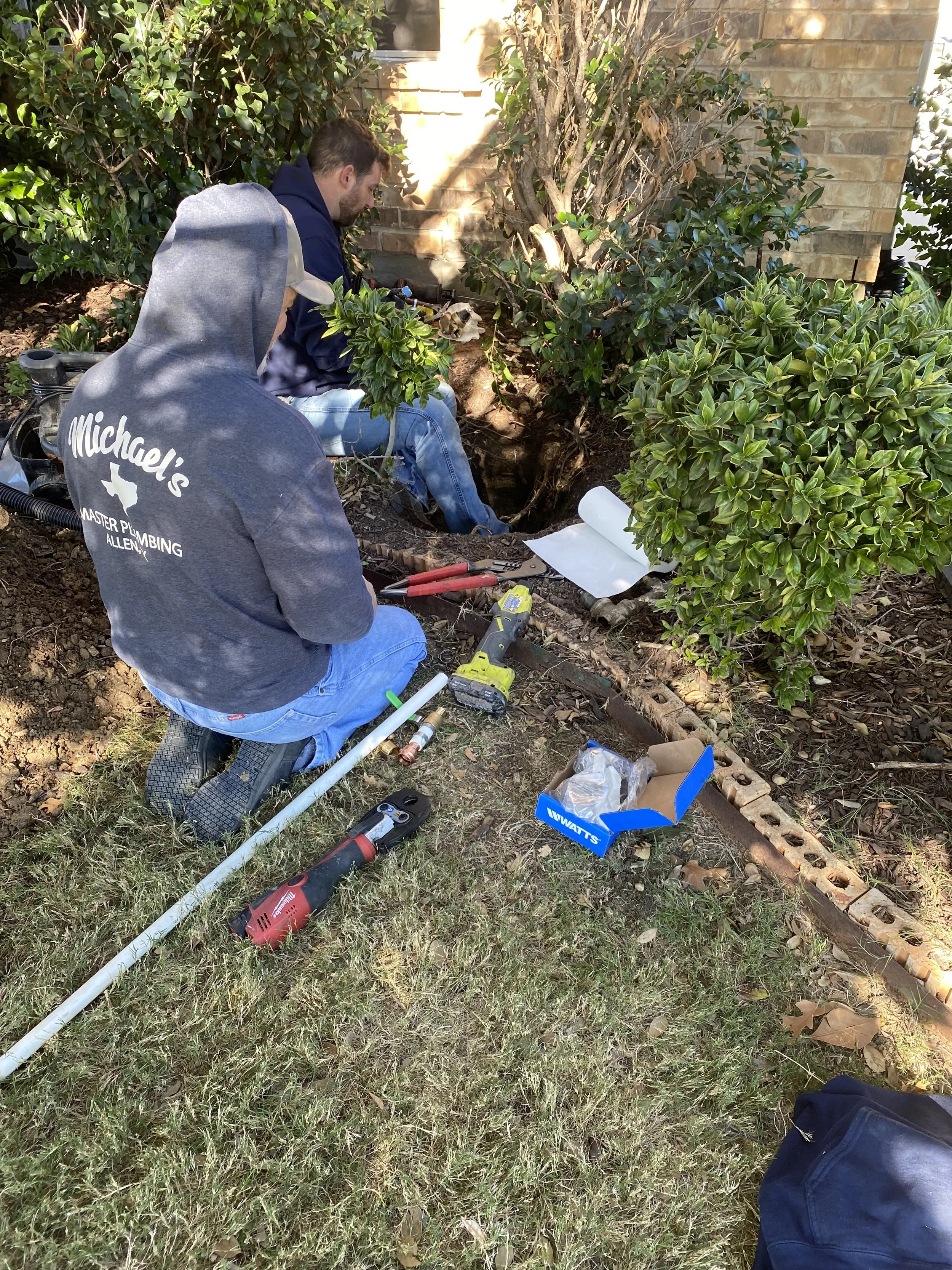 Two workers are digging in a garden area near a brick house, surrounded by bushes and small trees. One worker is kneeling with tools nearby, and the other is sitting and working on the ground.
