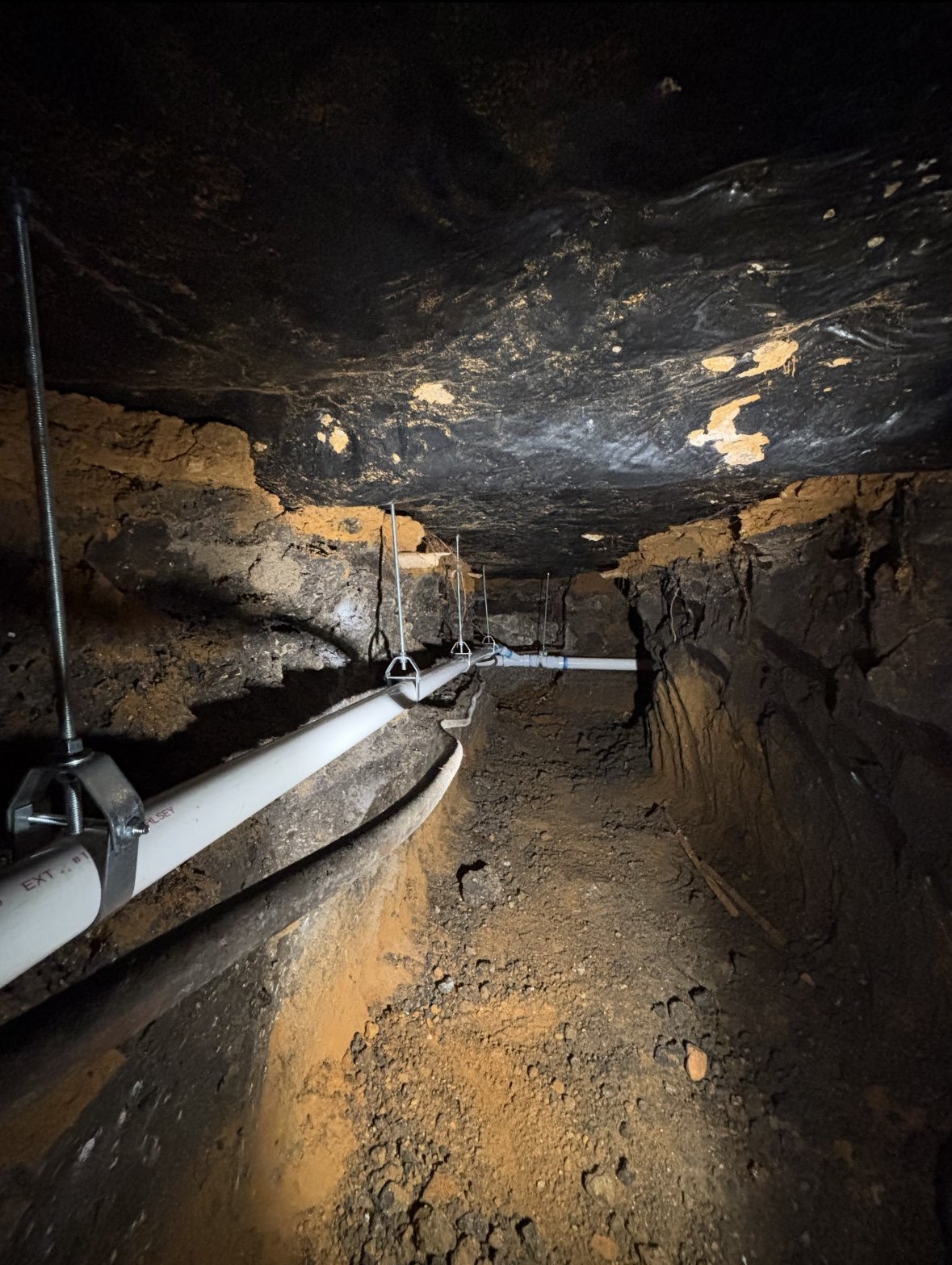 Underground tunnel with exposed rock walls and ceiling, containing a white pipe fastened with metal clamps, running along the left side of the tunnel.