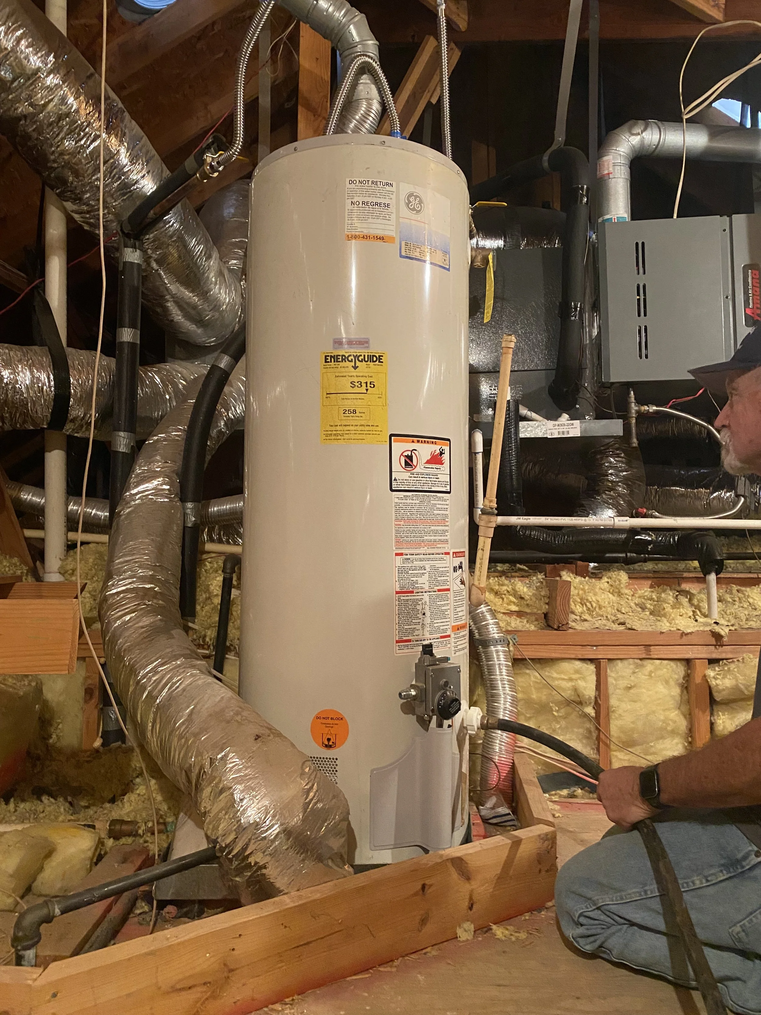 A tall, cylindrical water heater in a basement or utility room, with various ducts, pipes, and cables connected to it. A person is kneeling nearby, possibly inspecting or servicing the unit.