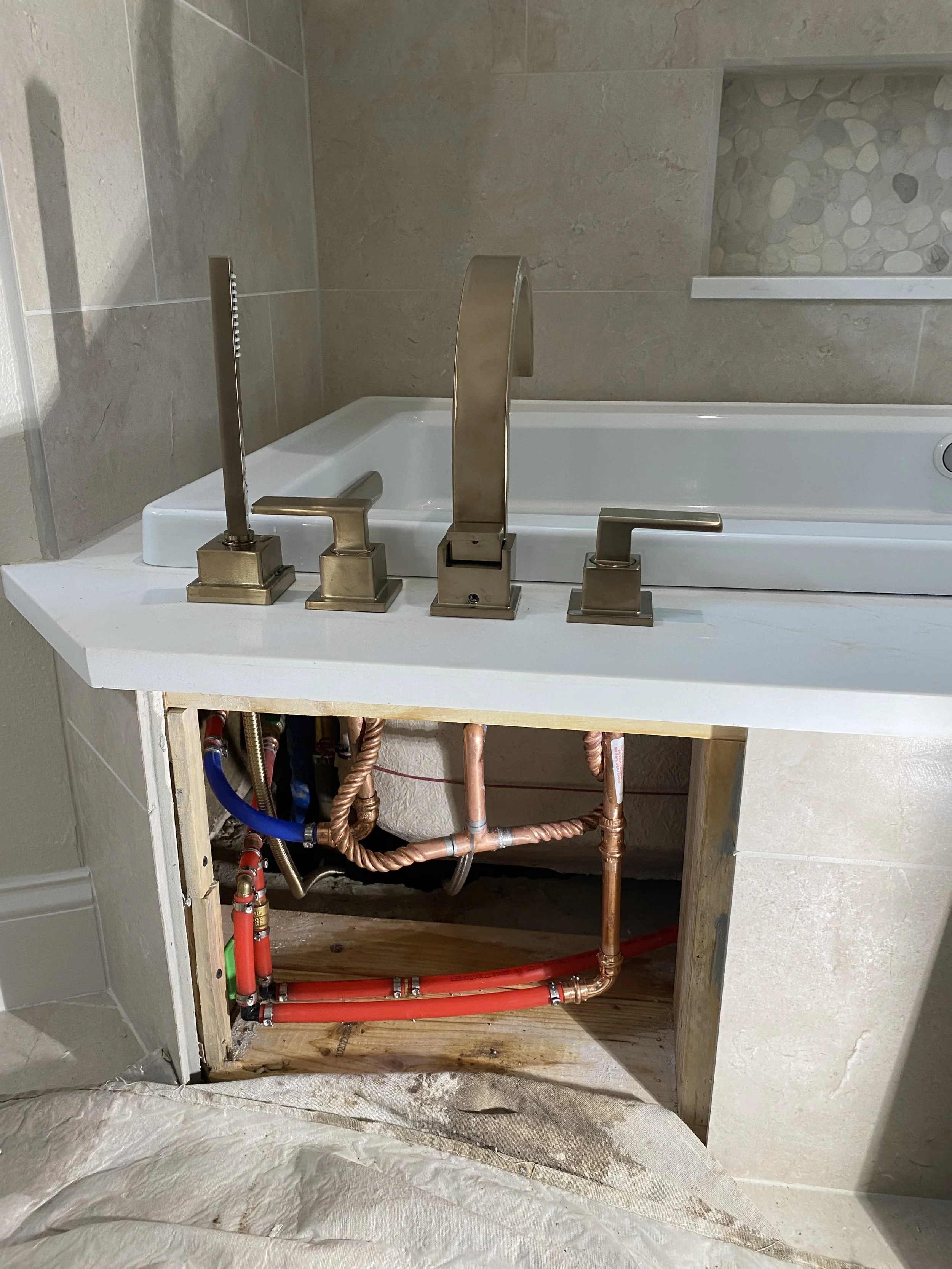 Bathroom sink with visible plumbing underneath, including copper pipes, red PEX pipes, and water supply lines, with a partially installed white countertop and a bathtub in the background.