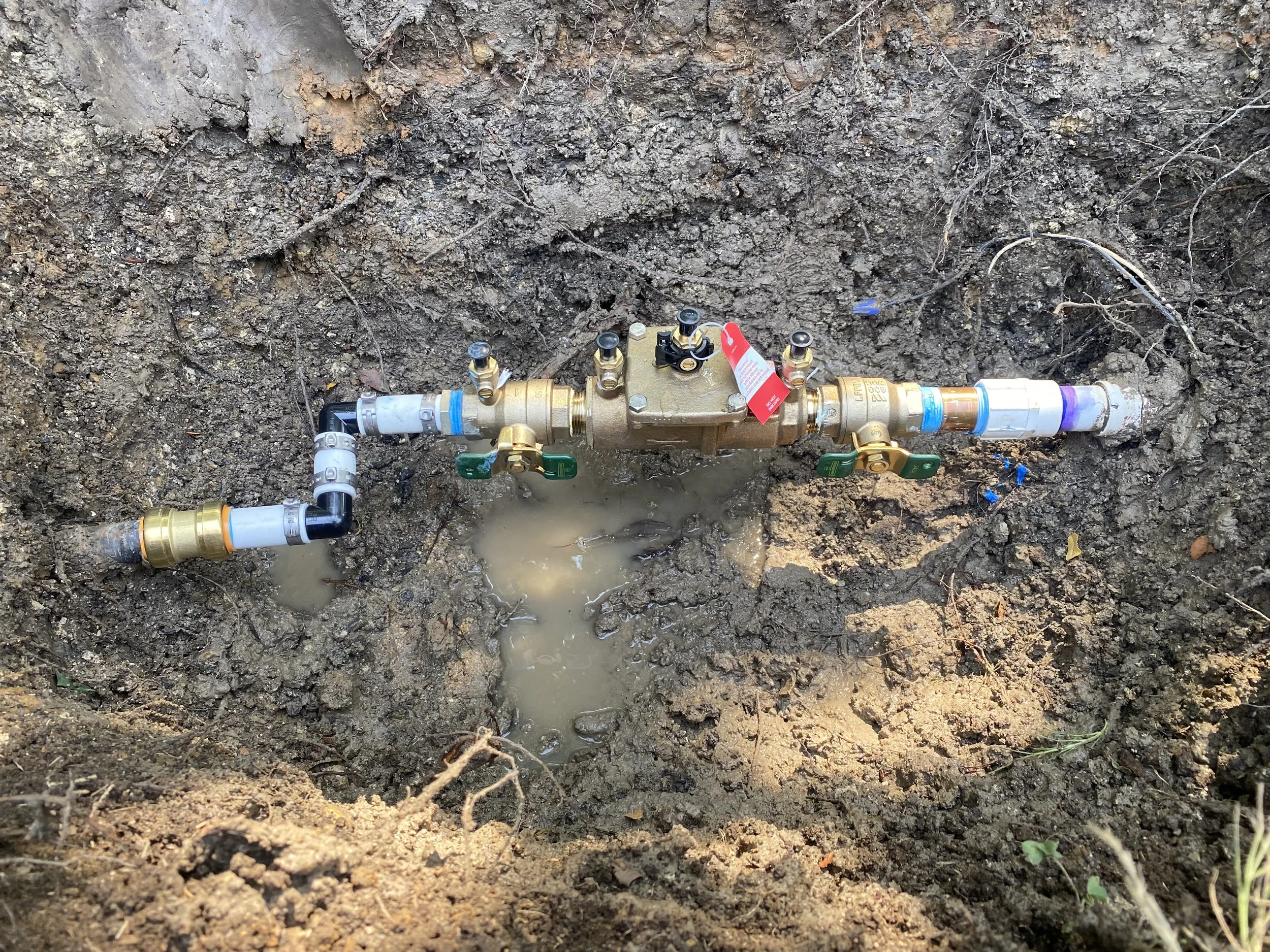 A buried water valve with pipe connections, valves, and gauges installed in a hole in the ground, with some water pooled around it.