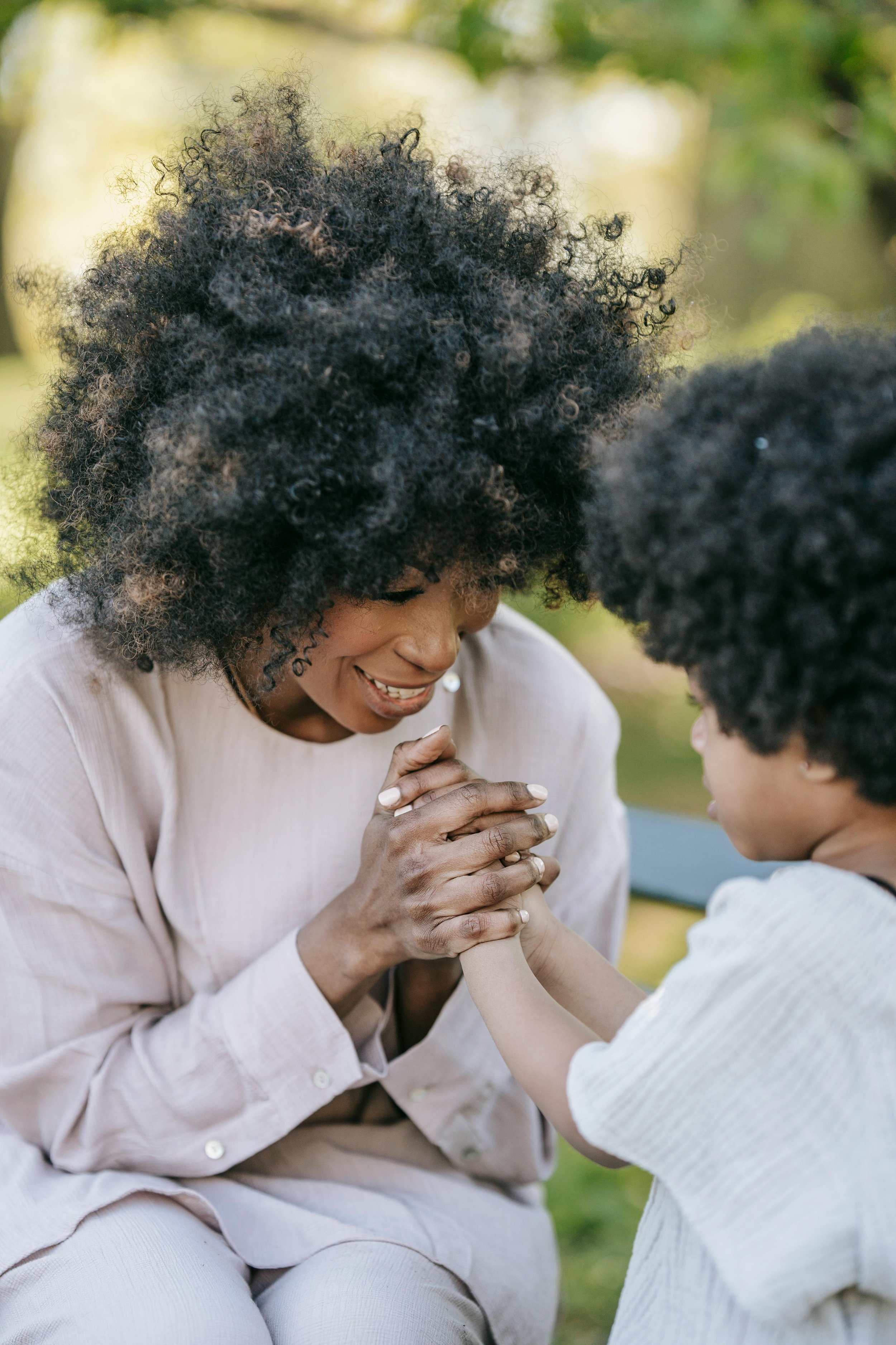 A woman and a child engaging in a tender moment outdoors, with the woman smiling and clasping the child's hand in a gesture of connection and affection, surrounded by blurred greenery.