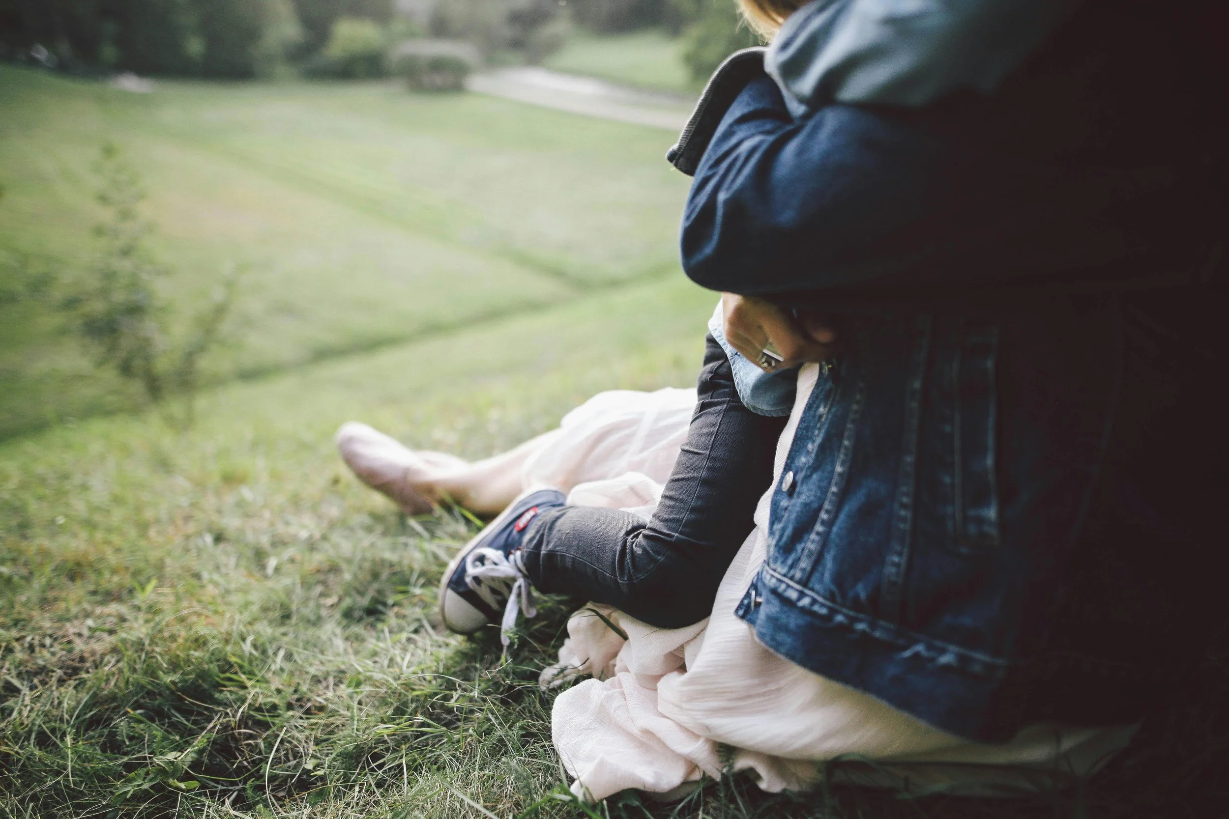 Person sitting on grass in a field with another person lying next to them, and a third person partly visible, background features greenery and trees.