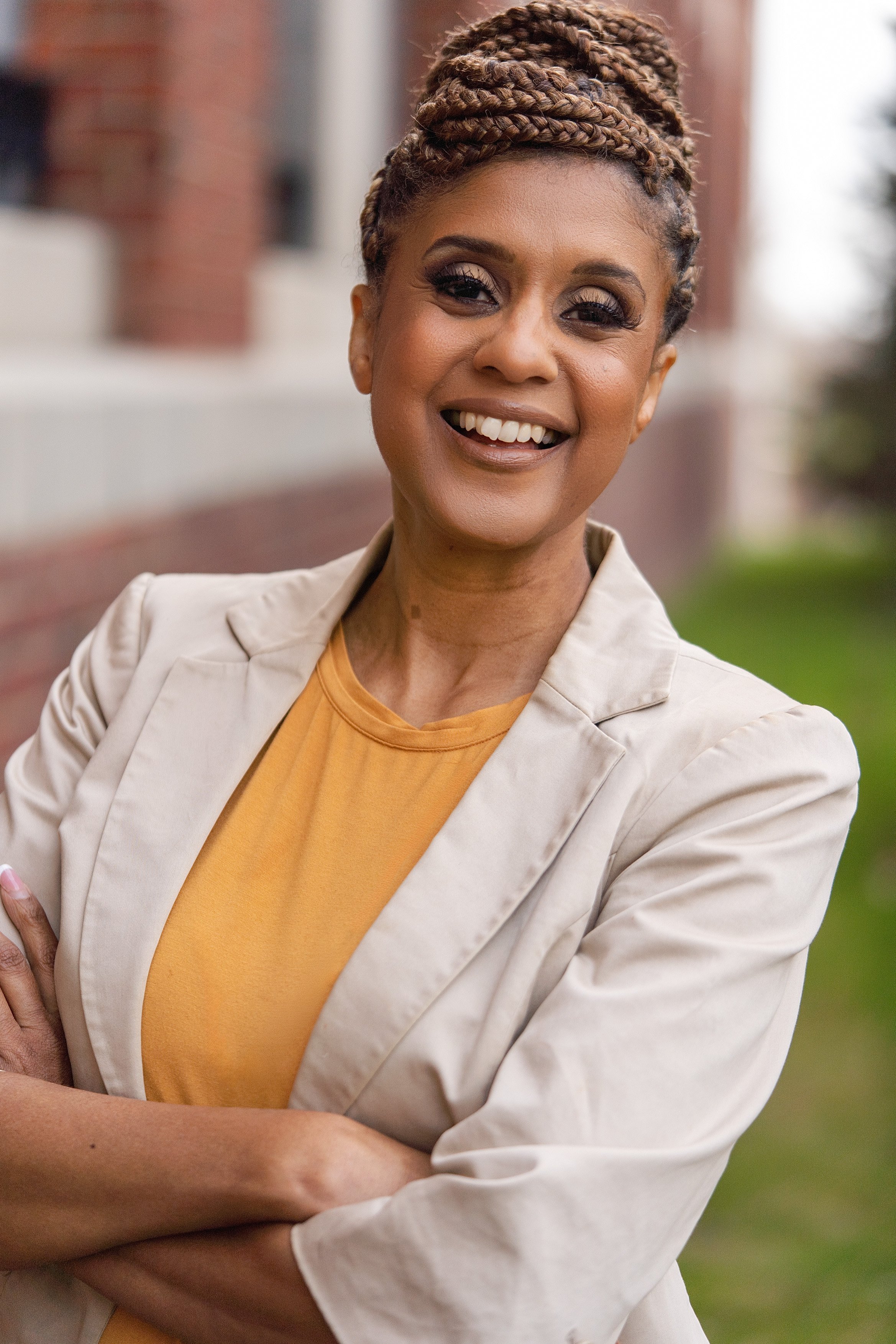 Evelyn Fordham Goodman smiling with braided hair wearing a beige blazer and yellow shirt, posing outdoors with arms crossed.