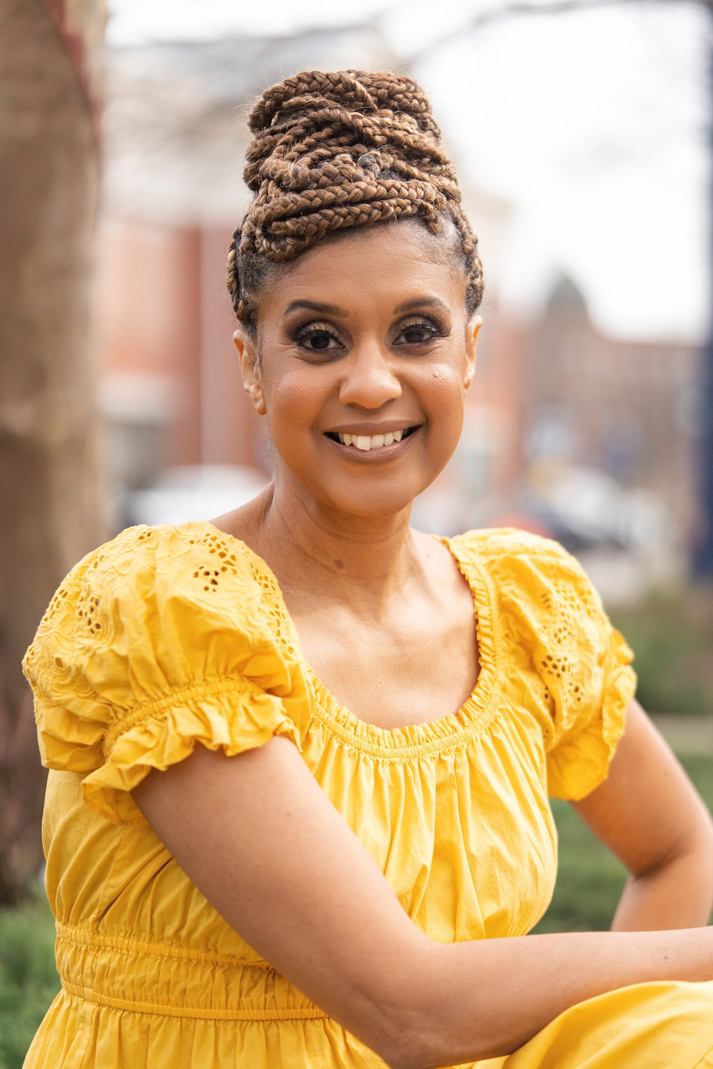 Evelyn Fordham Goodman with styled braids in a high bun, smiling, wearing a yellow dress with puffed sleeves, outdoors in a park-like setting.
