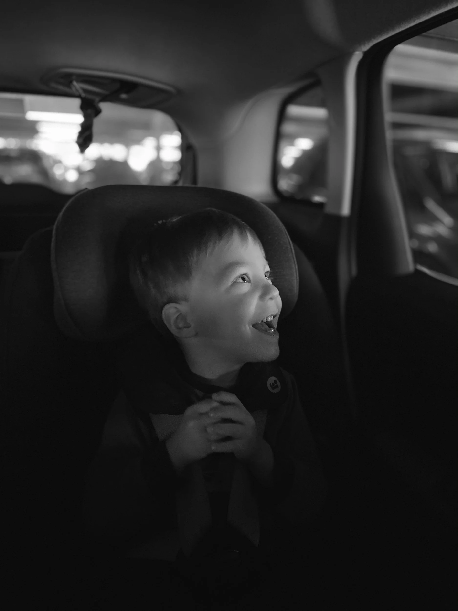 A young boy sits in a car seat inside a vehicle, joyfully looking upward with a big smile, hands clasped together, in a parking garage.