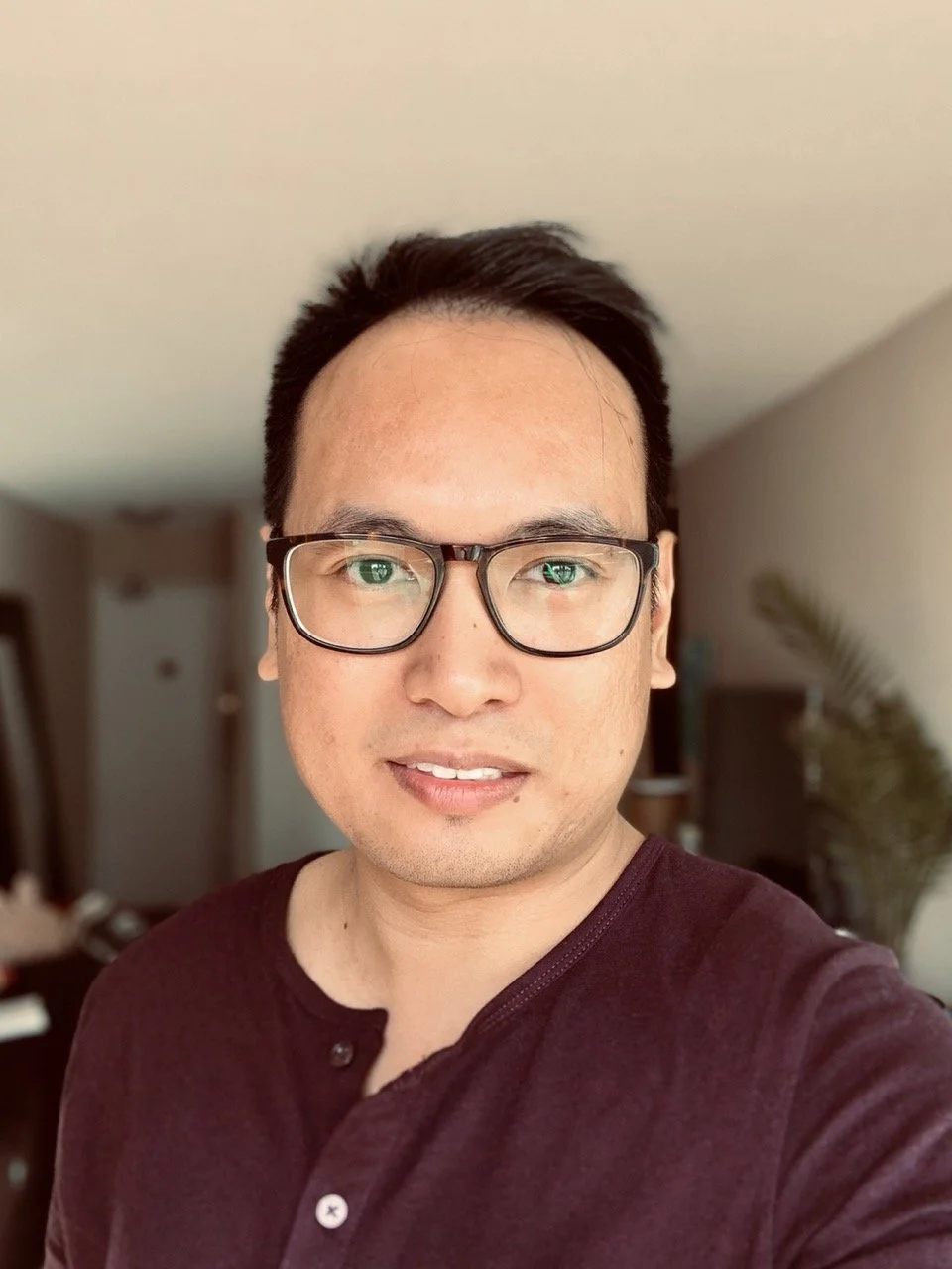 A young man with dark hair and glasses smiling in a room with a blurred background, including a plant and furniture.