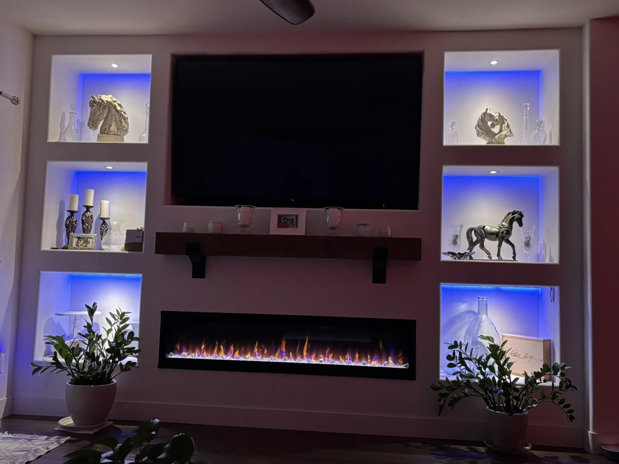 A living room with a modern electric fireplace, built-in shelves with decorative items, vases, candles, and plants, illuminated with blue and warm lighting.