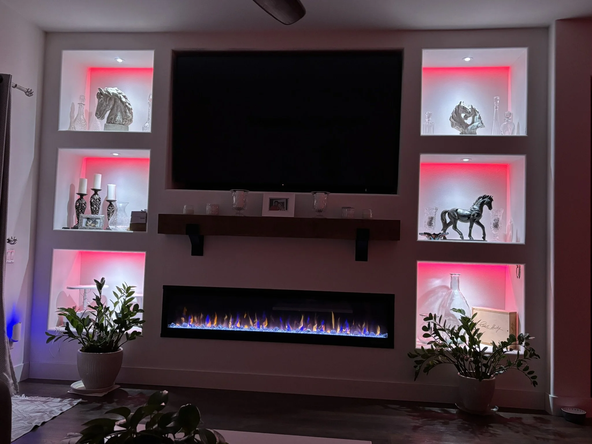 Living room with a wall-mounted flat-screen TV surrounded by illuminated shelves displaying decorative items such as sculptures, vases, and candles. The fireplace below the TV has colorful flames, and there are potted plants on the floor in front of 