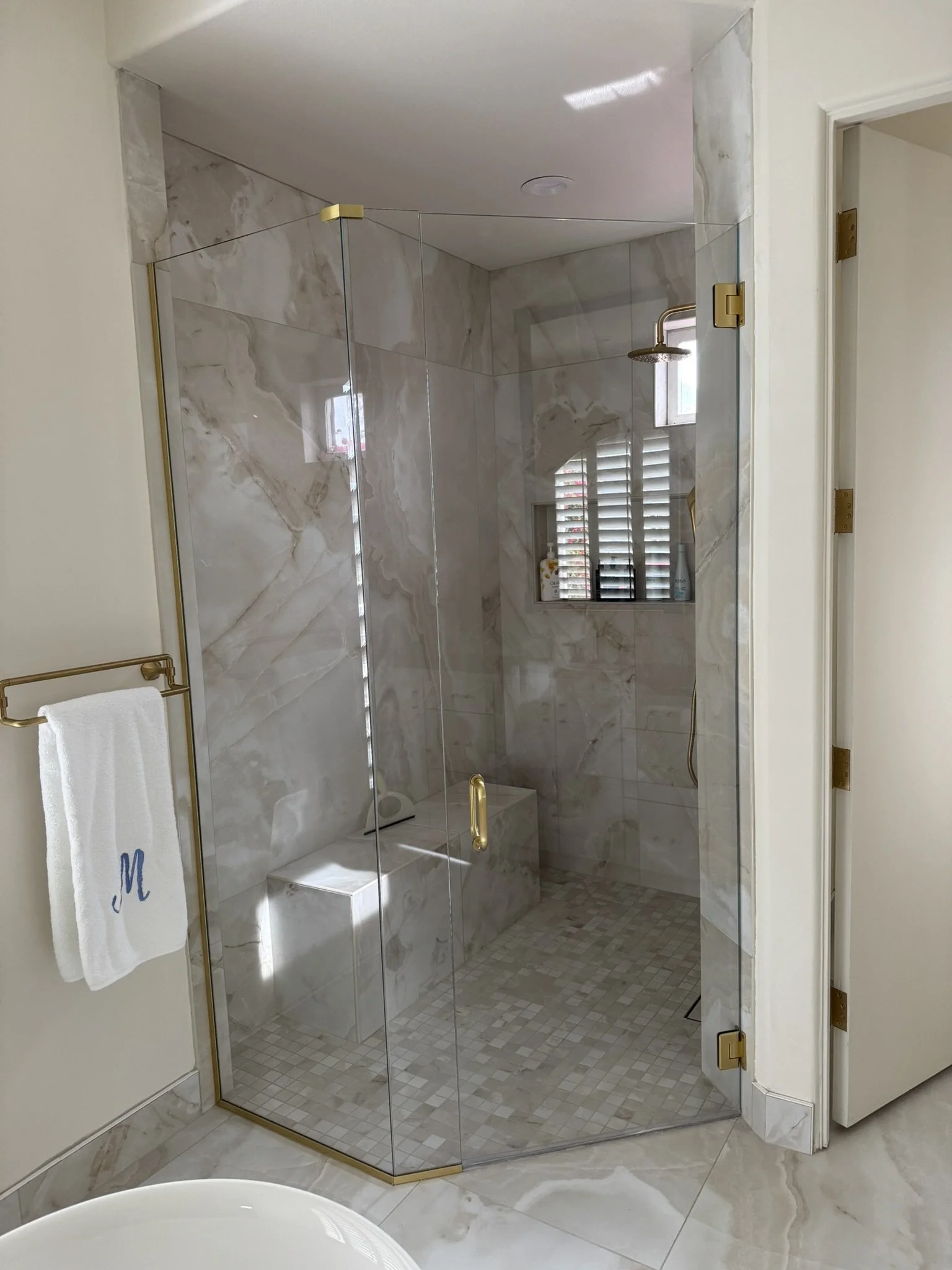 A walk-in shower with glass door and gold fixtures, marble walls, a window with shutters, and a built-in bench inside a modern bathroom.