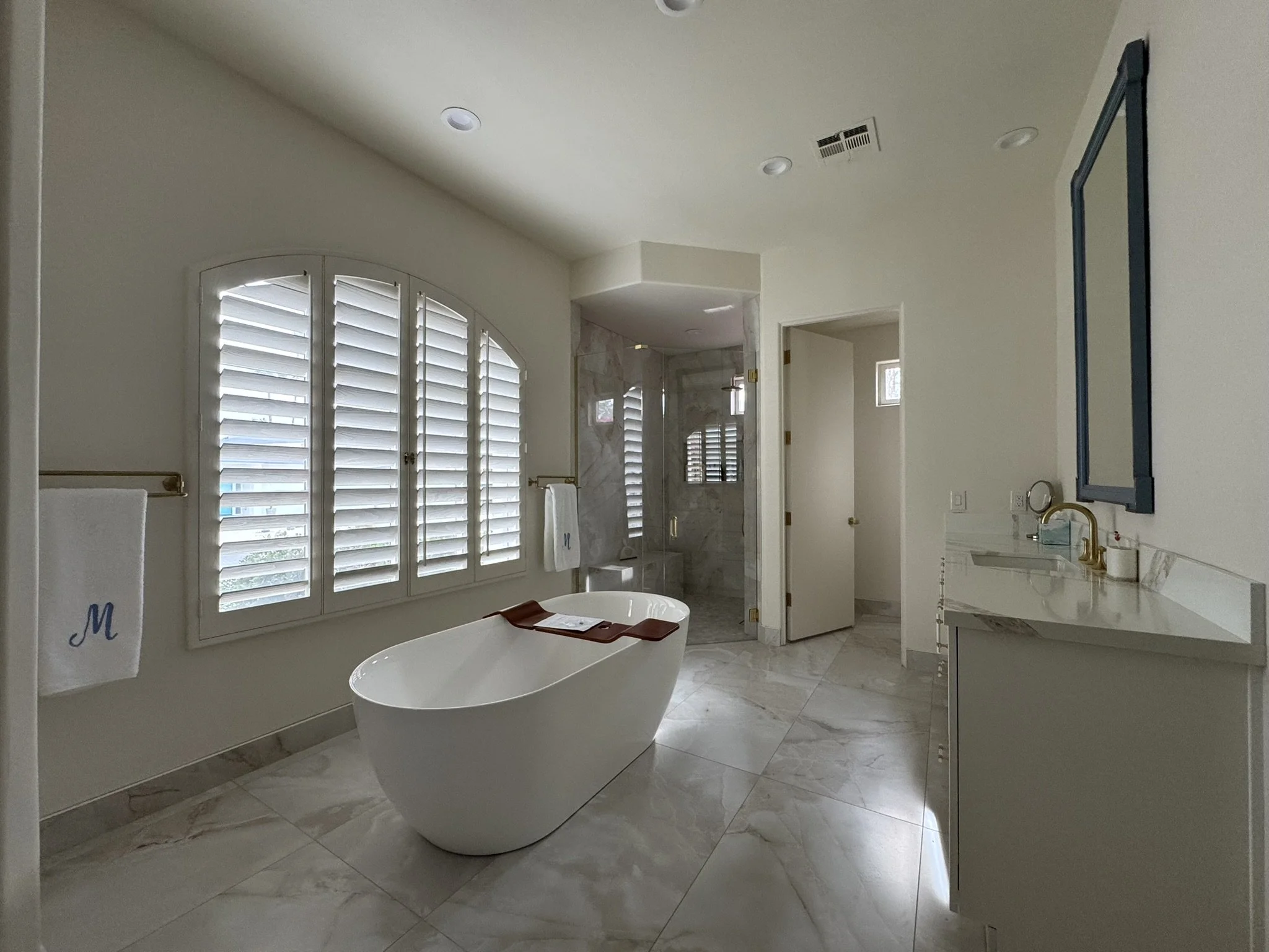 Luxurious bathroom featuring a free-standing oval bathtub, large marble tiles, a window with white plantation shutters, a vanity with gold fixtures, and a walk-in shower with a glass door.