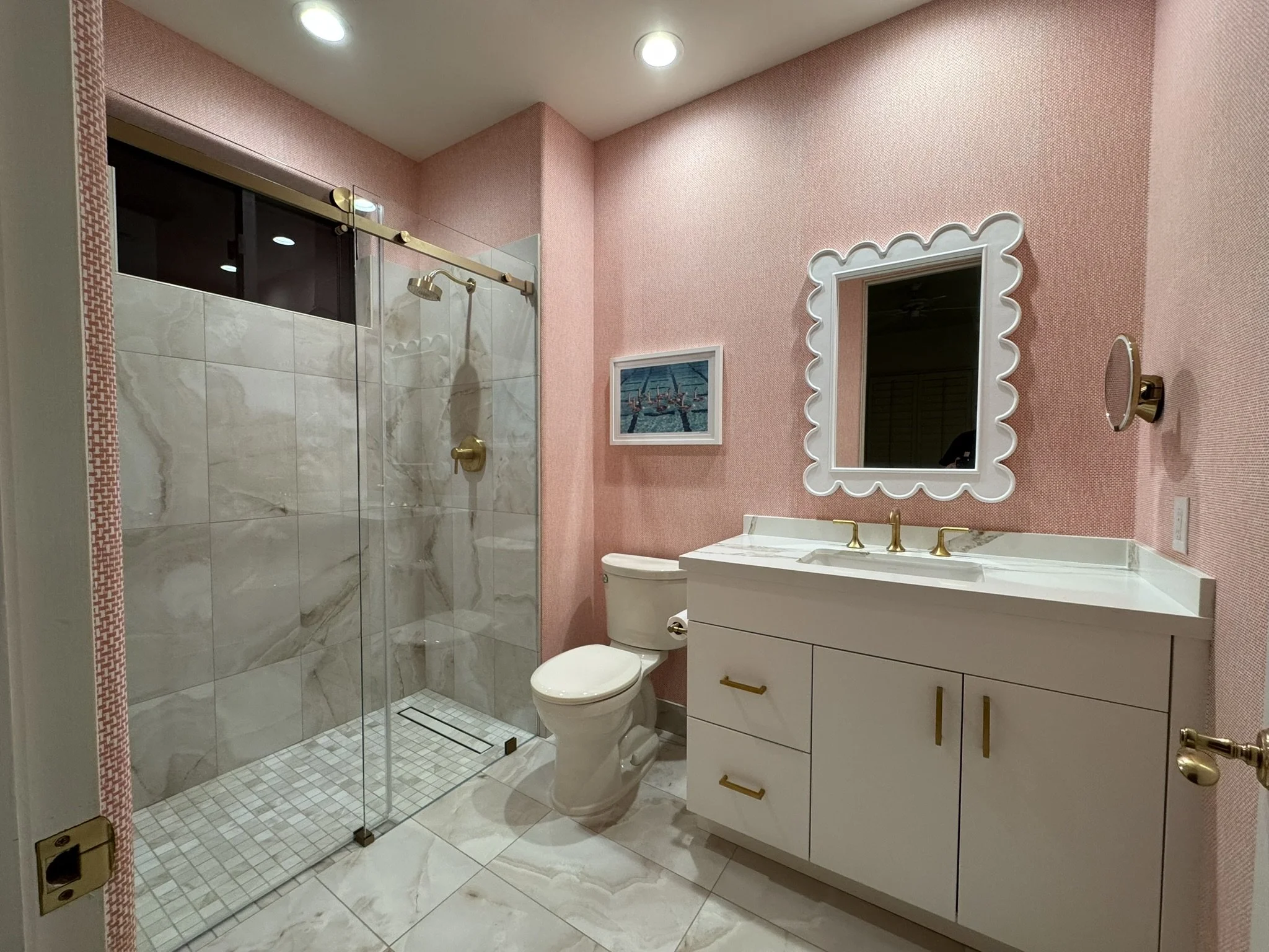 A bathroom with pink textured walls, a white vanity with gold hardware, a mirror with a scalloped frame, a toilet, and a separate glass-enclosed shower with marble tiles and a gold shower head. There's a framed picture on the wall and recessed lighti