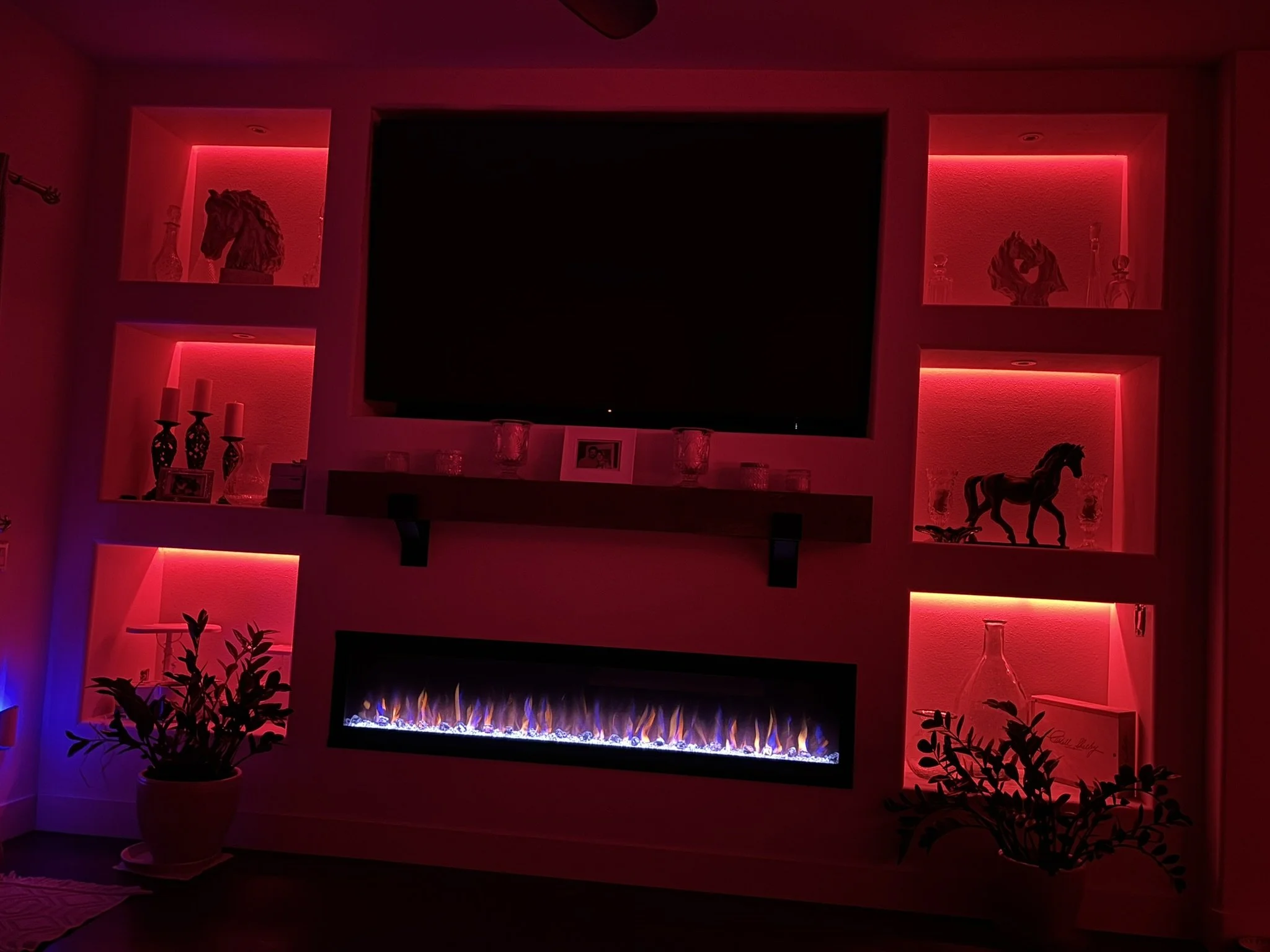Living room with a TV above a fireplace, illuminated by red and blue LED lights, with decorative items on shelves and potted plants in the foreground.
