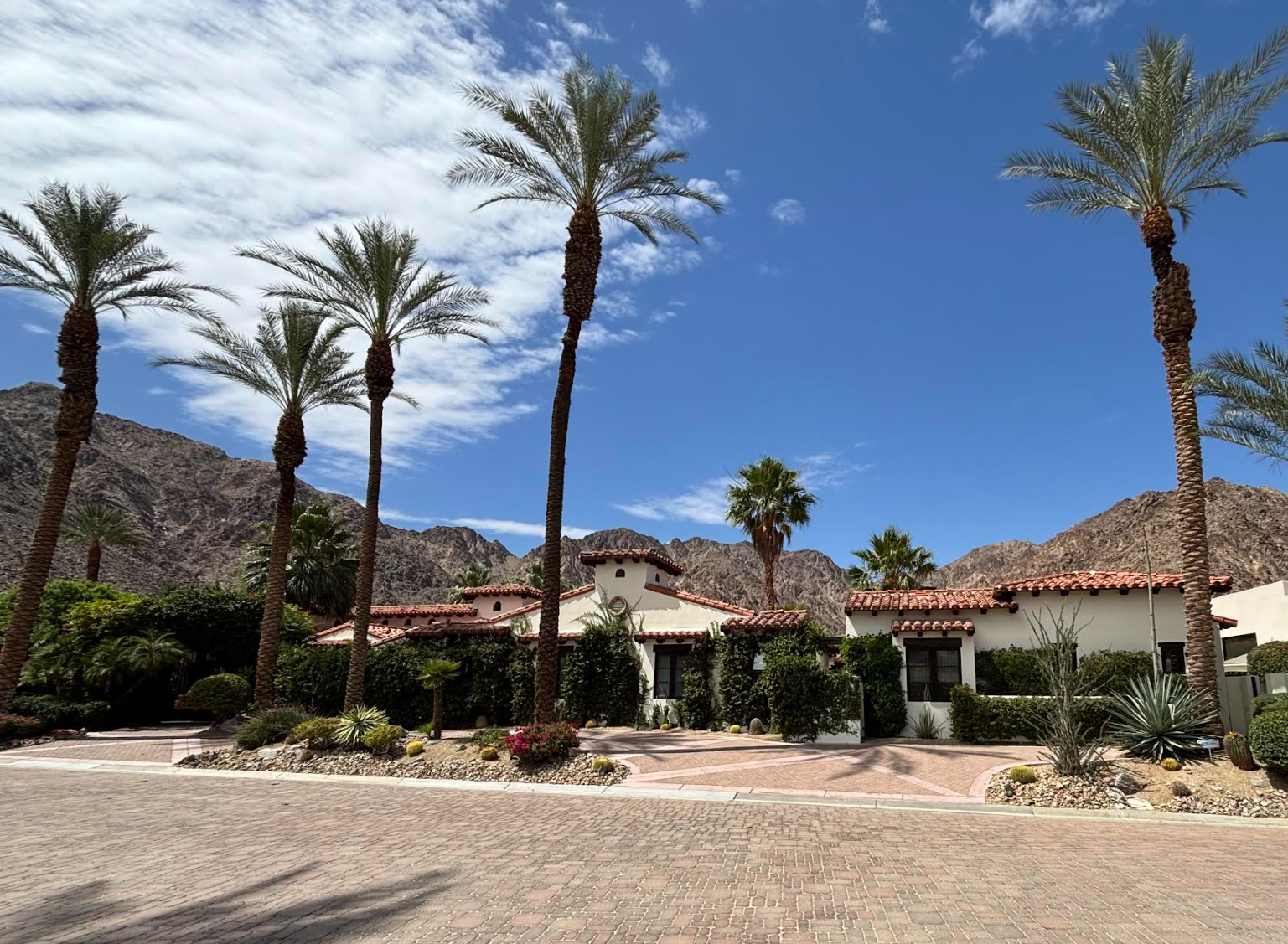 A house with Spanish-style architecture surrounded by palm trees and desert mountains under a partly cloudy sky.