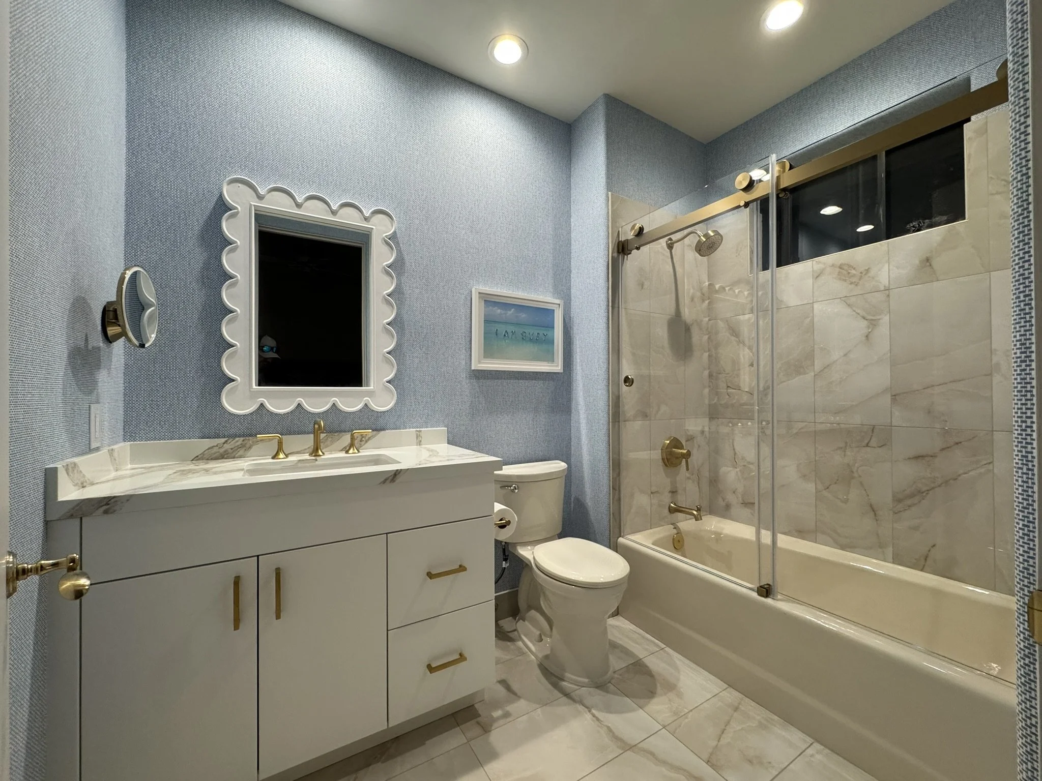 Modern bathroom with blue textured walls, white vanity with gold hardware, framed mirror, toilet, bathtub with glass door, and marble tiles.