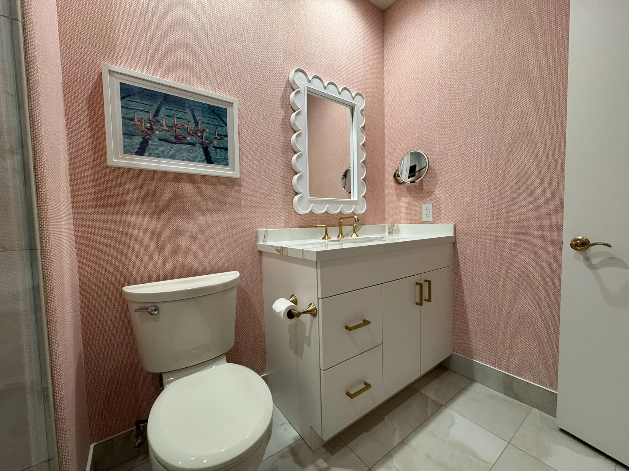 A small bathroom with pink textured wallpaper, a white toilet, a white vanity with gold hardware, a decorative framed mirror, a small round magnifying mirror on the wall, and a colorful framed art piece of ballet dancers near a swimming pool.