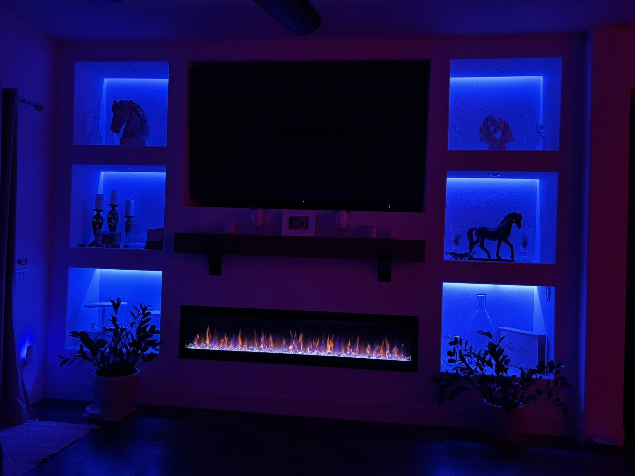 Living room with a modern electric fireplace, illuminated display shelves with decorative items, and potted plants, all bathed in blue and purple ambient lighting.