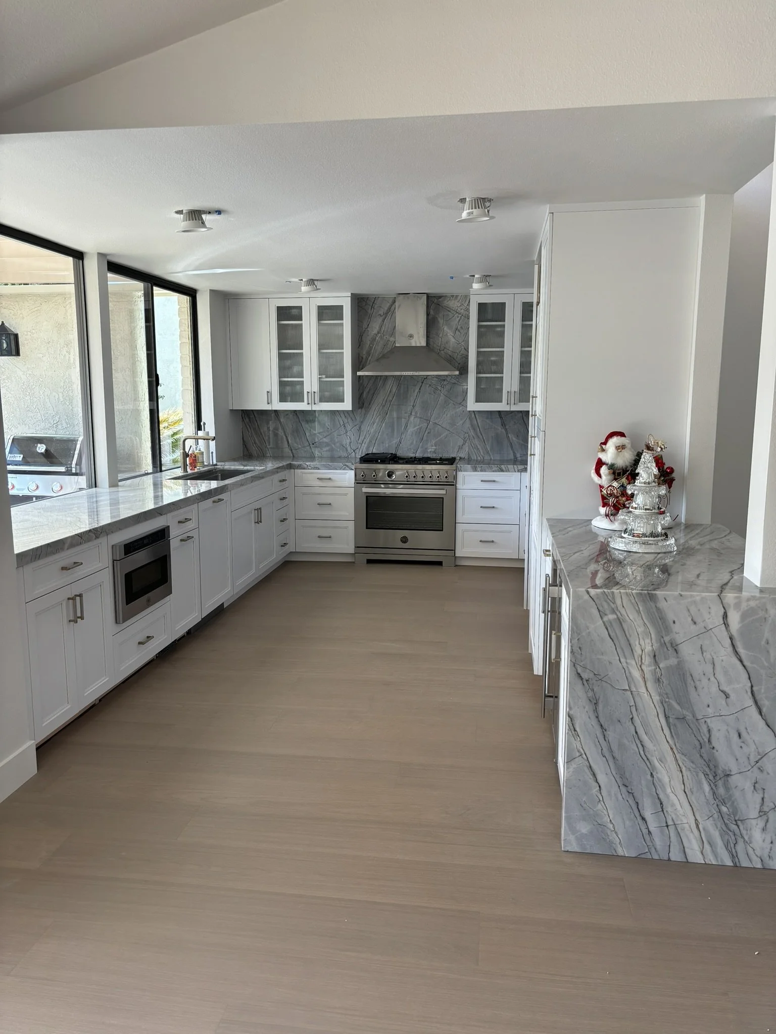 View of a modern kitchen with white cabinets, marble countertops, a stainless steel stove and oven, a marble backsplash, and a window showing an outdoor patio with a grill. Christmas decorations, including a Santa figurine and a glass tiered tray wit