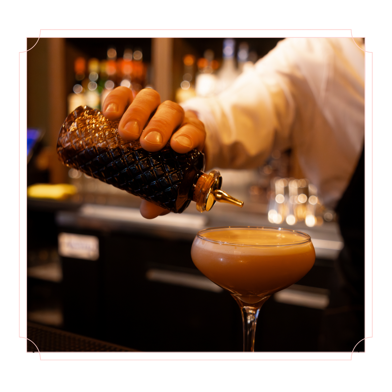 Bartender pouring a drink from a syrup bottle into a cocktail glass at a bar.