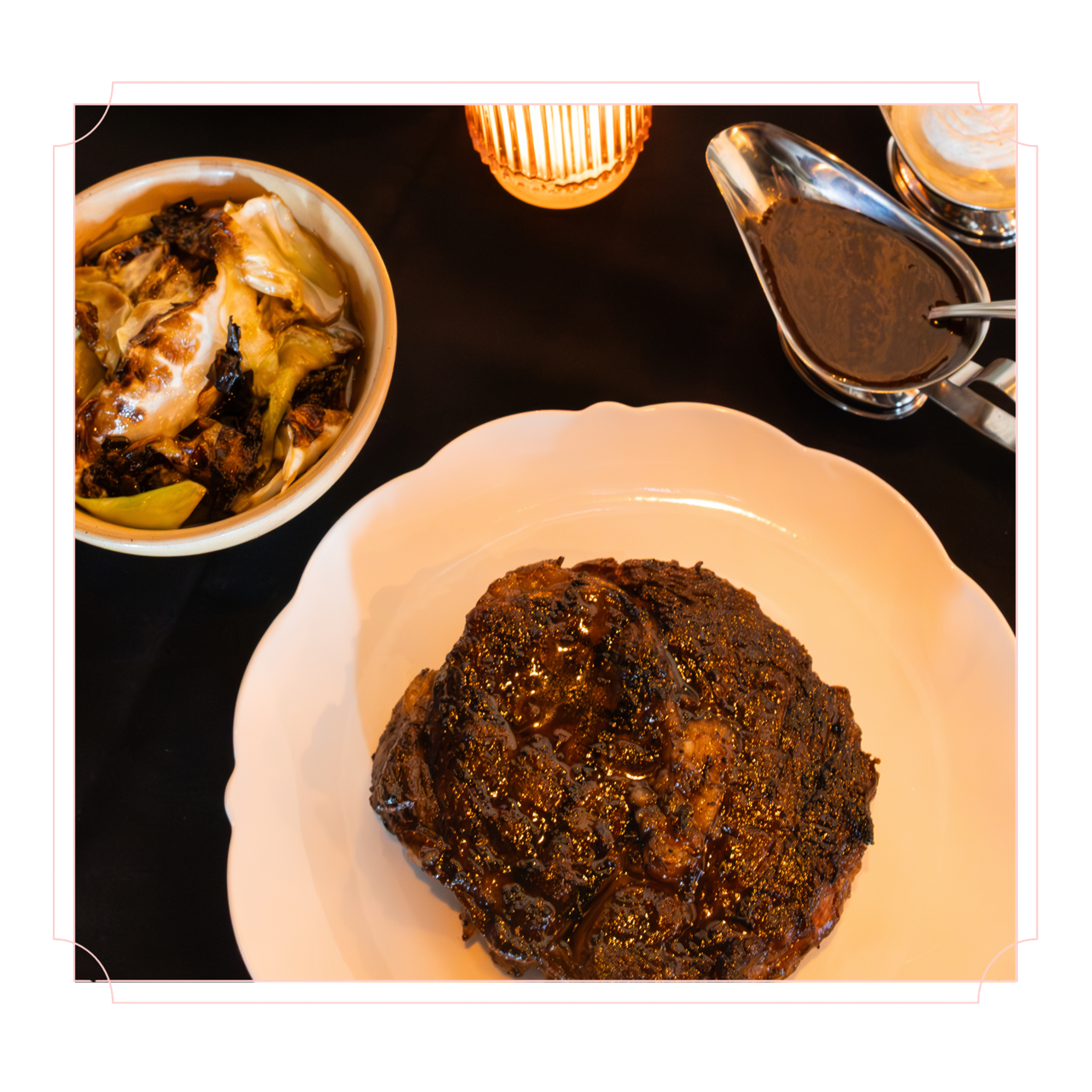 A large, cooked piece of meat with sauce on a white plate, a bowl of cooked vegetables, a small pitcher of gravy, and a small container of salt on a black table, with a lit candle in the background.