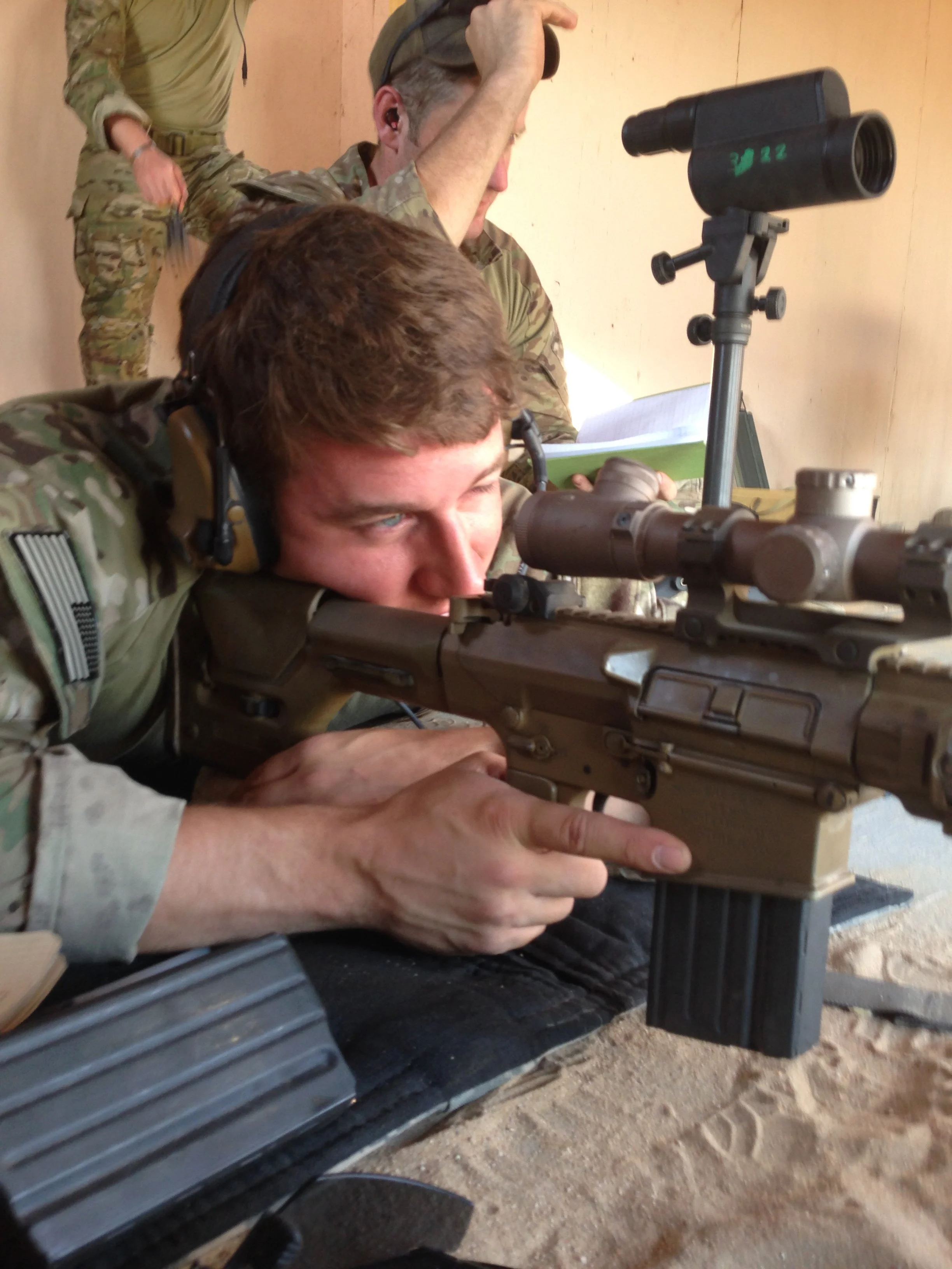 A green beret aiming a sniper rifle through a scope, lying on the ground with other soldiers in the background.