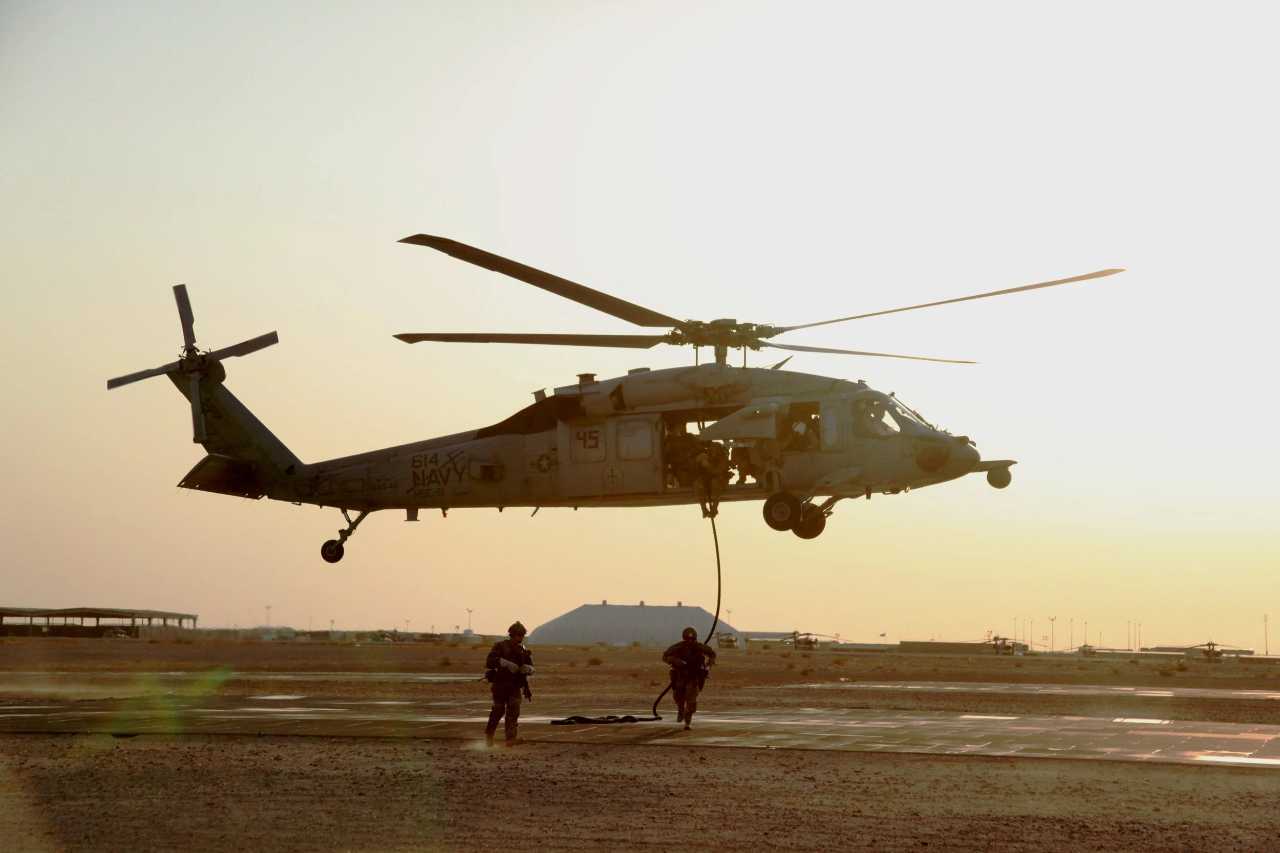 Military helicopter hovering above ground while several special forces members fast rope down and provide security, in a desert-like environment during sunset