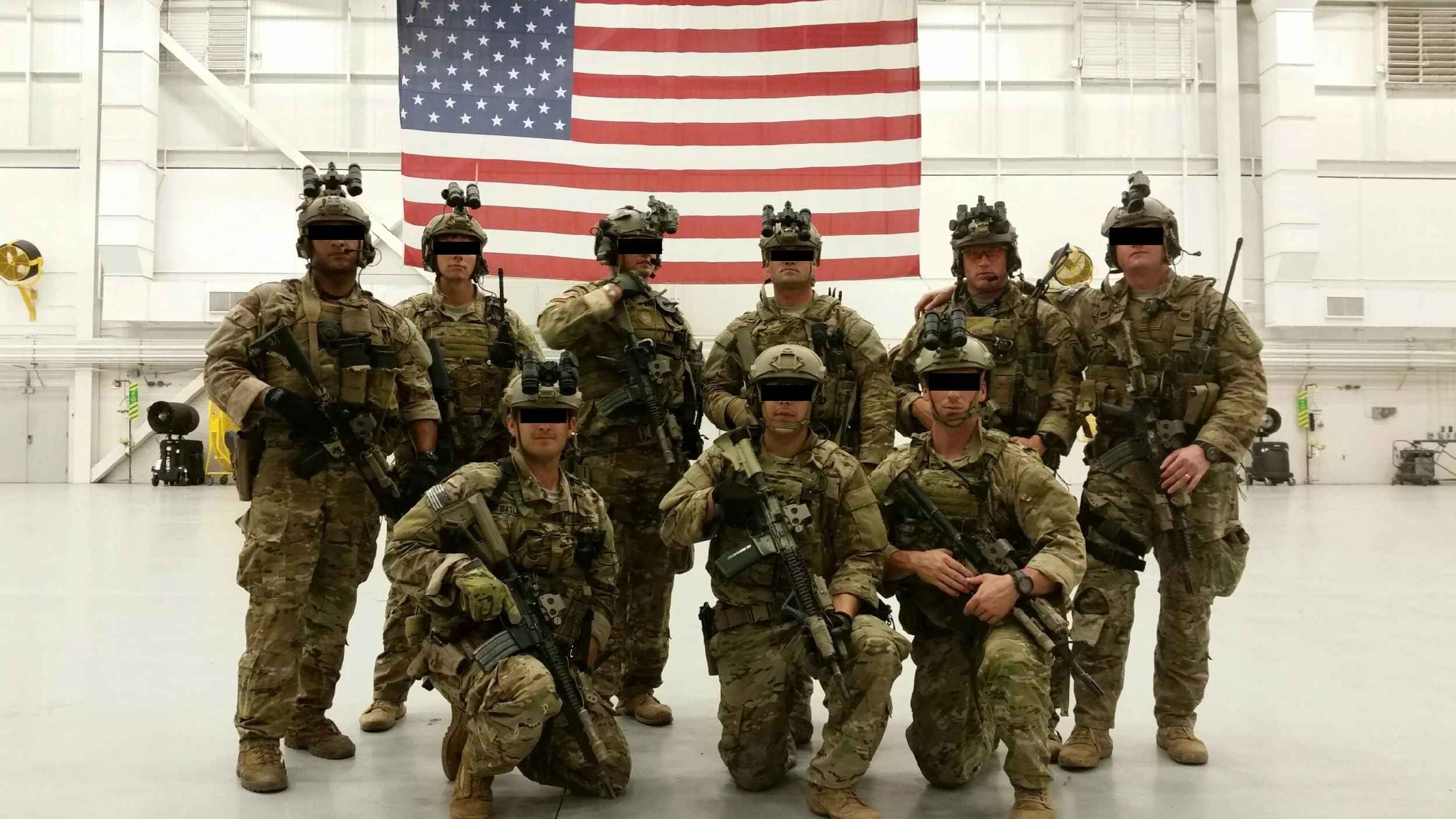 A group of nine special forces soldiers in combat gear inside a large hangar with an American flag hanging on the wall behind them.