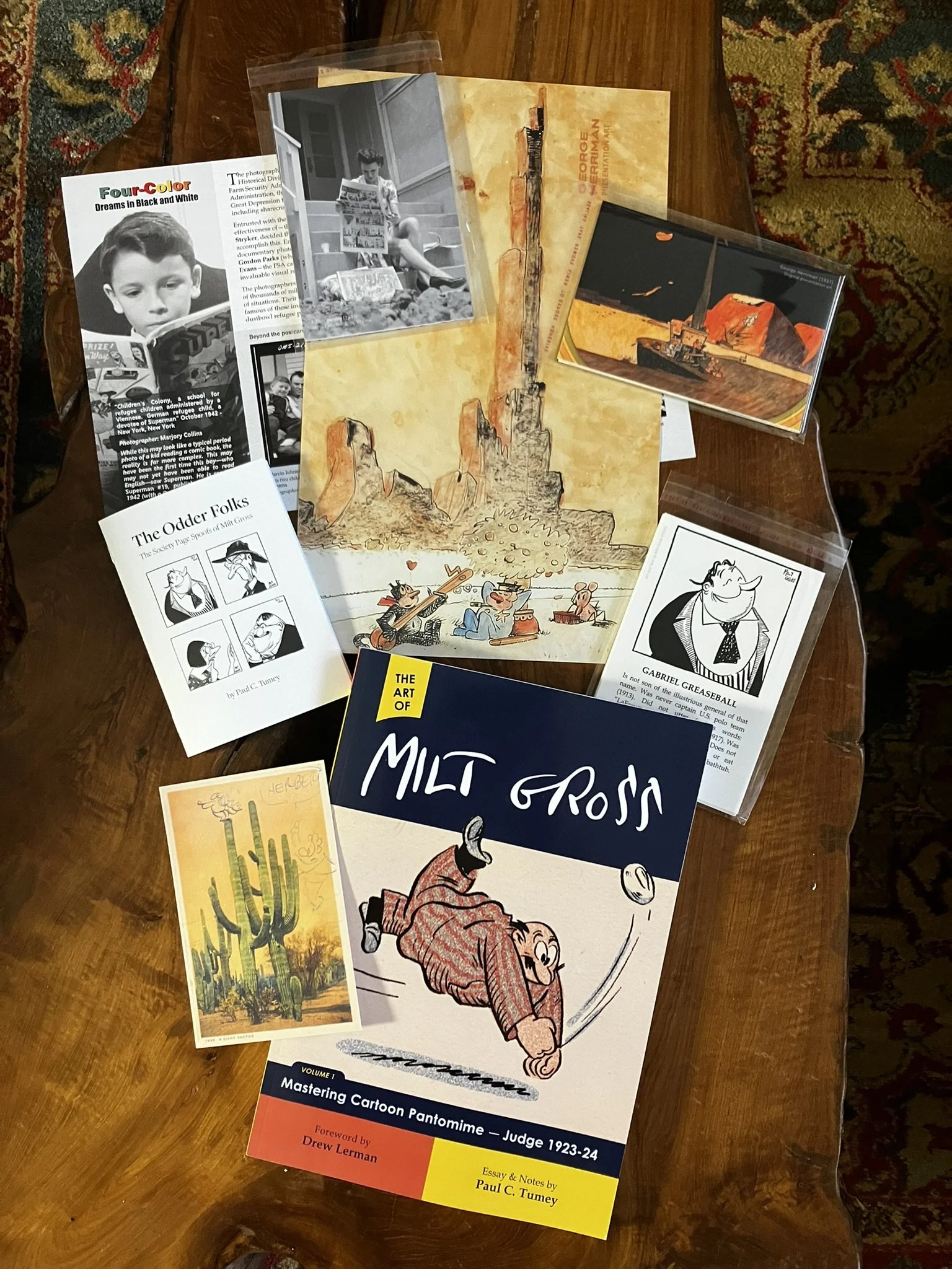 A table with multiple cartoon and historical art books, illustrations, black-and-white photographs, and notes arranged on it.