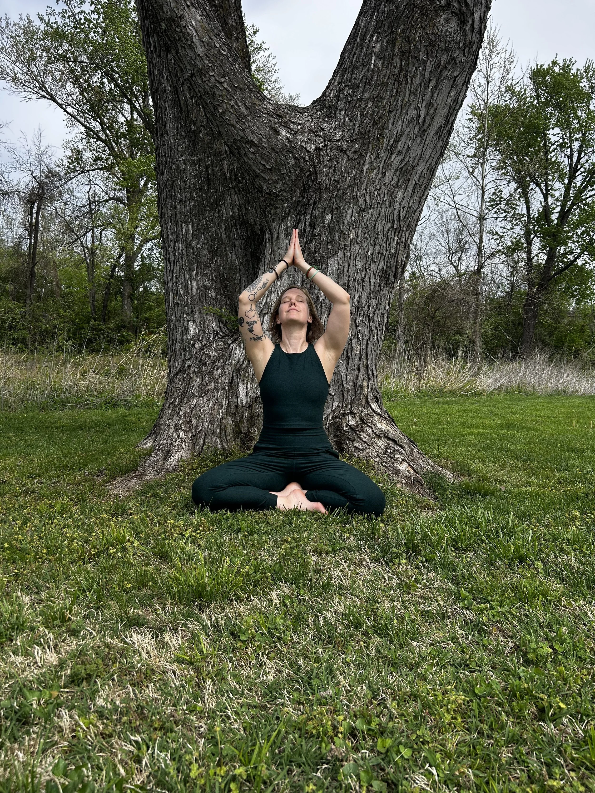 A woman practicing yoga outdoors in a seated meditation pose with hands in prayer position above her head, sitting on grass in front of a large tree, surrounded by trees and greenery.