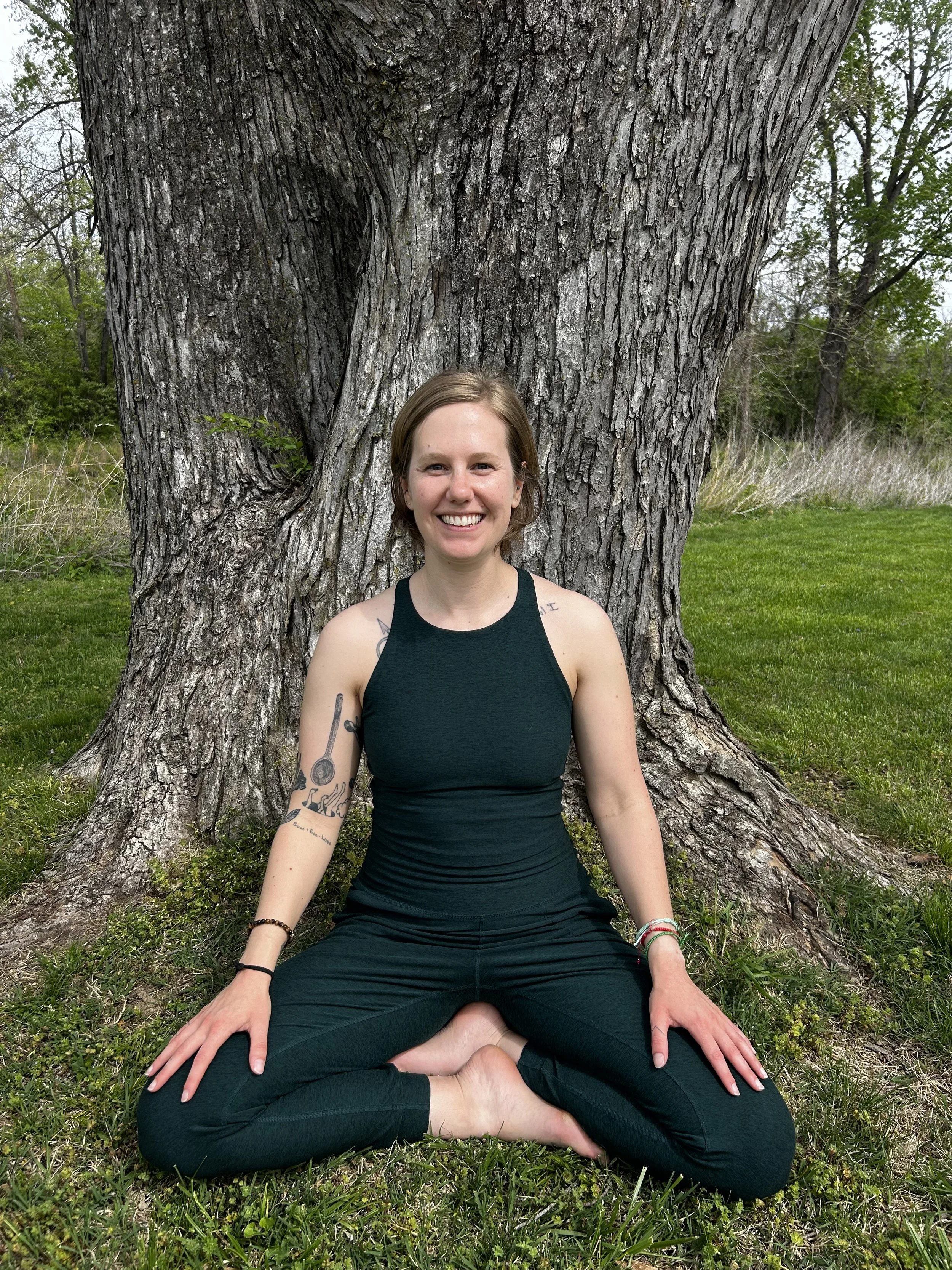 A woman with short blond hair, wearing a green tank top and green pants, sitting cross-legged on grass in front of a large tree with textured bark, smiling at the camera.
